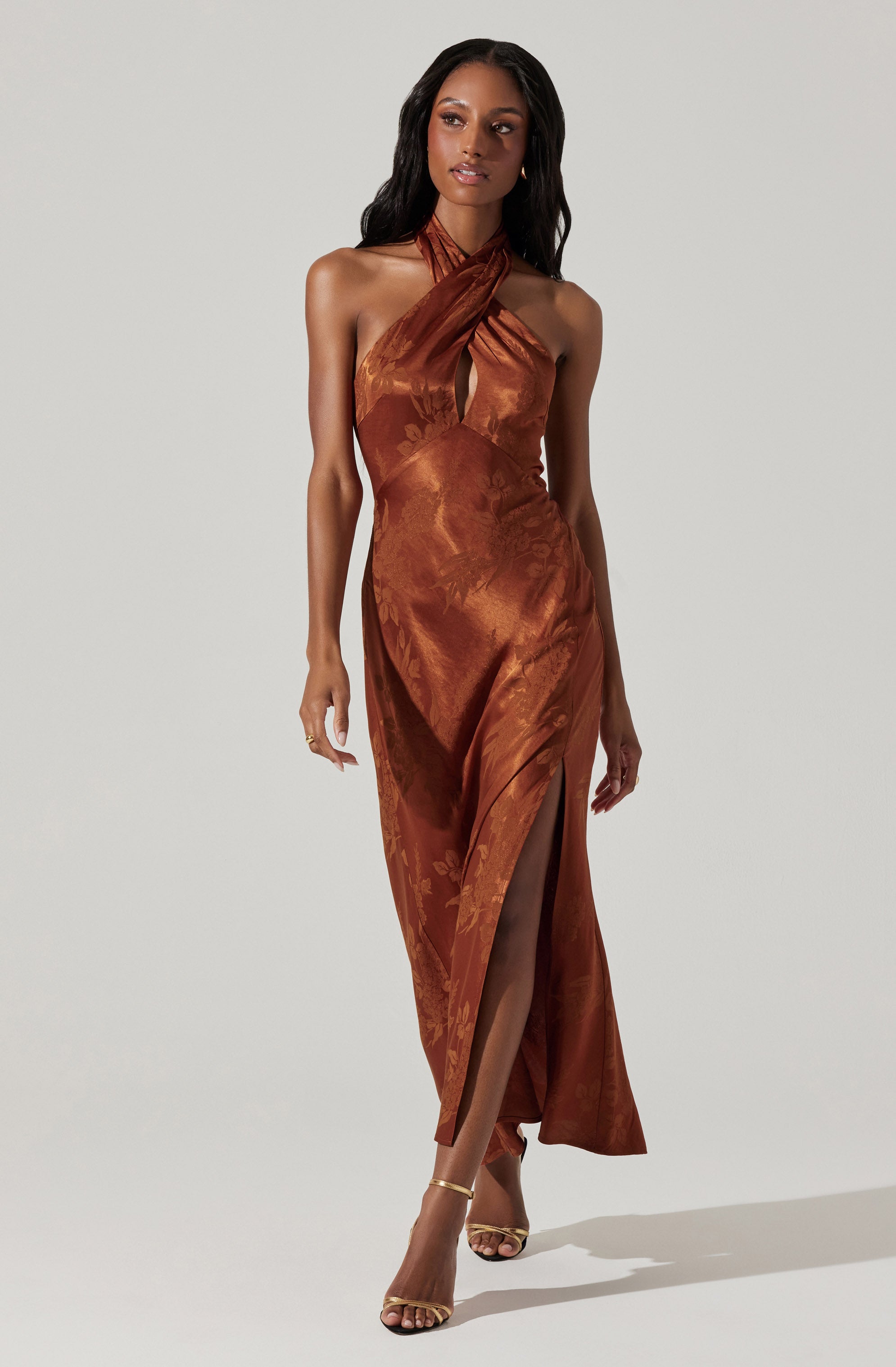 A woman is wearing a long, satin, rust-colored dress with a floral pattern. The dress features a halter neckline with fabric crossing over at the chest and a high slit up one leg. She is also wearing strappy, open-toed heels.
