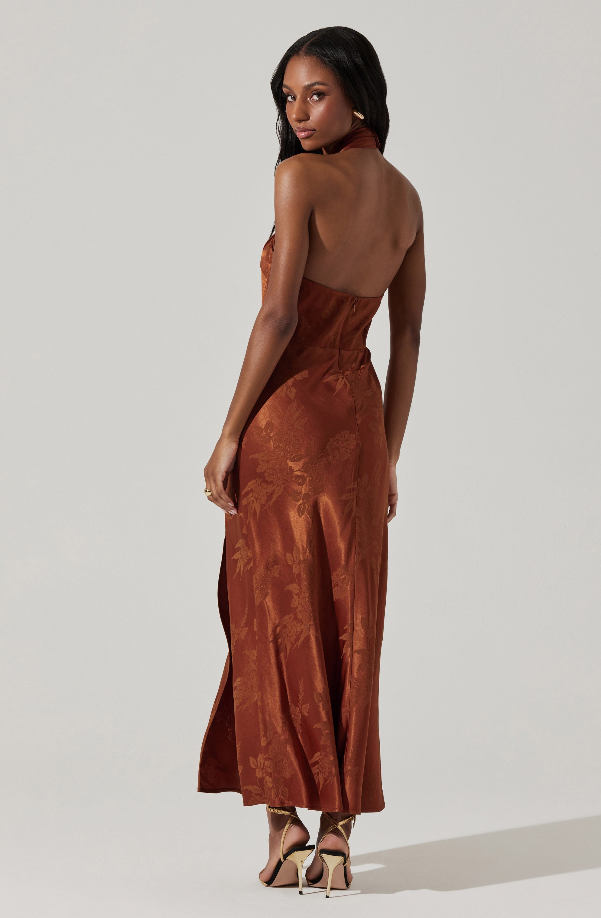 A woman is wearing a sleeveless, halter-neck, full-length gown in a rich, rust color with a subtle floral pattern. The dress appears to be made of a shiny, satin-like fabric. She is also wearing high-heeled sandals with ankle straps.