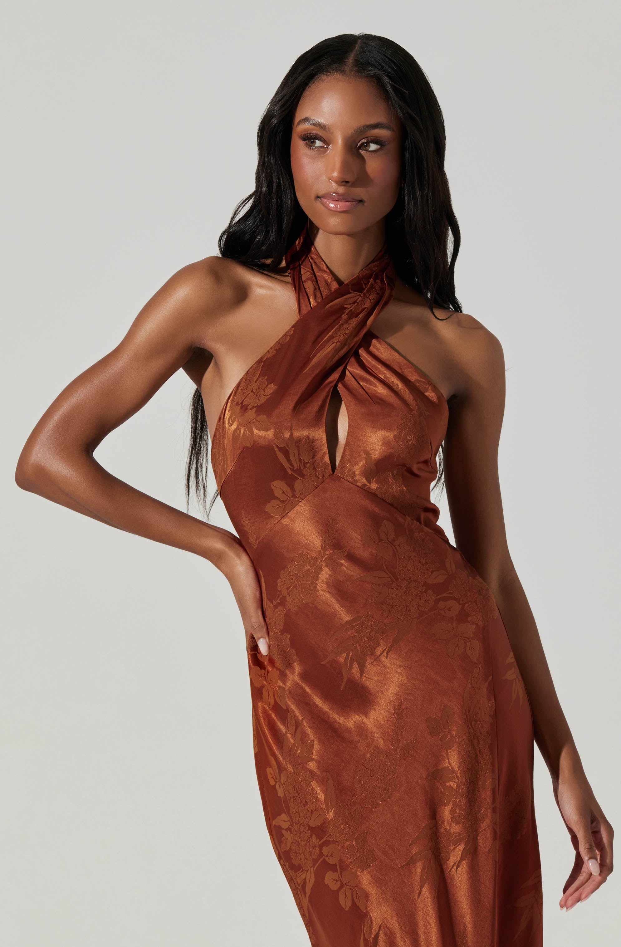 A woman is wearing a sleeveless, satin-like dress in a rich, burnt orange color. The dress features a halter neck design with a twisted fabric detail at the neckline, creating a slight keyhole opening. The fabric has a subtle floral pattern that adds texture and elegance to the overall look.