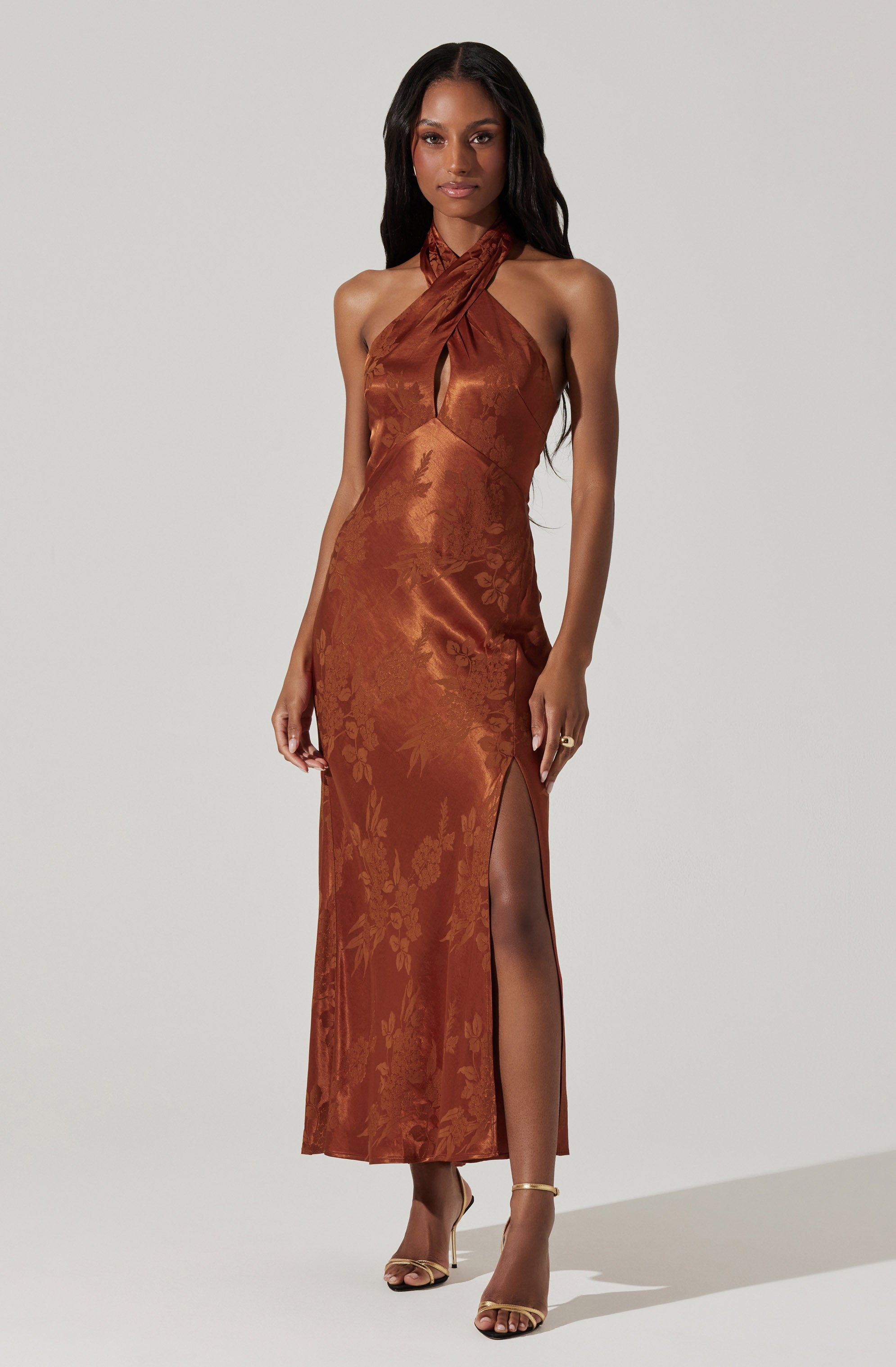 A woman is wearing a full-length, sleeveless, satin dress in a warm copper-brown color. The dress features a halter-style top with a twist at the neckline, creating a keyhole cutout at the chest. The fabric has a subtle floral pattern woven into it. The dress has a high slit on one side, revealing her leg, and she is wearing strappy high-heeled sandals.