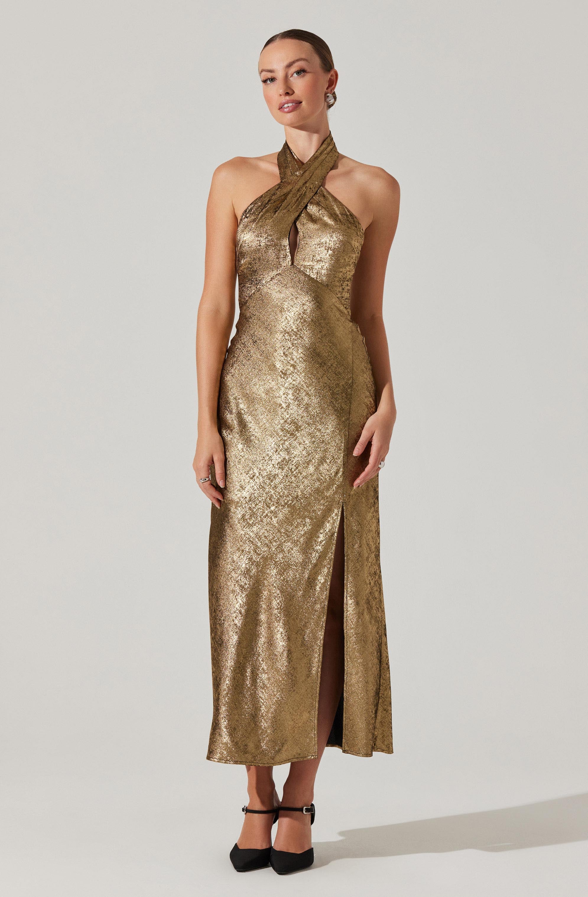 A woman is wearing a long, gold metallic gown with a halter neckline that features a twist front and a keyhole cutout at the bust. The dress has a high slit in the front, revealing her legs, and she is paired with black ankle-strap heels.