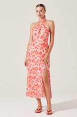 The dress is a sleeveless, full-length gown with a halter neckline that ties around the neck. It features a vibrant floral pattern in shades of pink and coral. The dress has a keyhole cutout at the chest and a high slit on one side, revealing the leg. The fabric appears lightweight and flowy. The model is accessorized with red strappy high-heeled sandals and gold earrings.