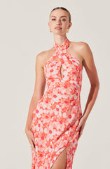 A woman is wearing a sleeveless, halter neck dress with a watercolor floral print in shades of pink, coral, and white. The dress features a keyhole cutout at the chest and a slit on the side, revealing part of her leg. She is accessorized with gold earrings.