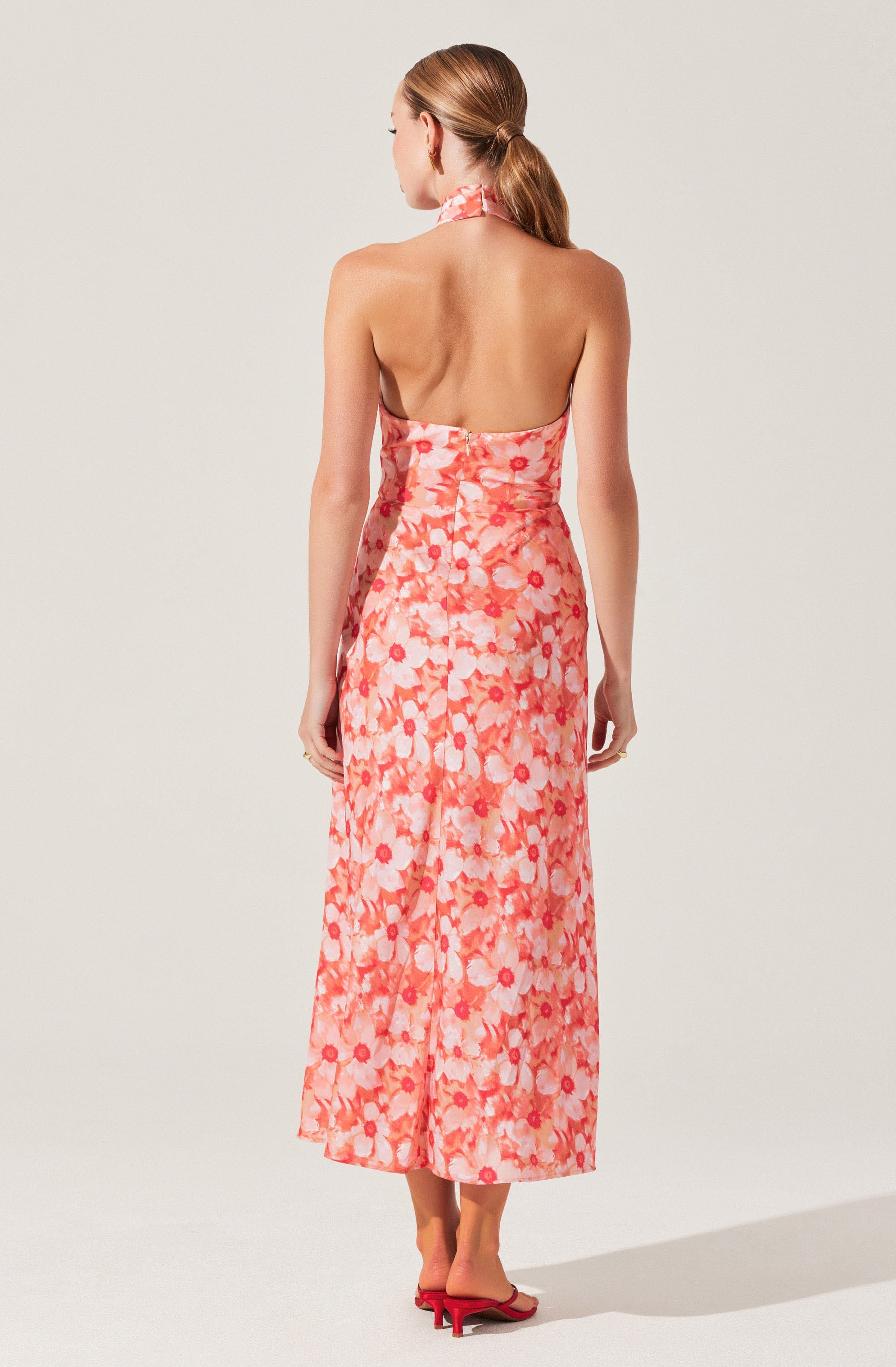 A woman is wearing a strapless, halter-neck dress with an all-over floral pattern in shades of pink, coral, and white. The dress has a fitted bodice and a long, flowing skirt that reaches her ankles. She is also wearing pink open-toe heeled sandals.