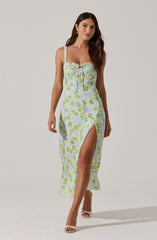 A woman is wearing a sleeveless, ankle-length dress with thin straps and a slit that reveals her leg. The dress has a light blue base color with a green and yellow floral pattern, and features a gathered, ruched bodice with a bow and tie detail at the center. She is also wearing white high-heeled sandals with an open toe.