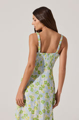 A woman is wearing a sleeveless dress with wide shoulder straps. The dress features a light background with a green and yellow floral pattern. The straps are gathered with a slightly ruffled detail. The dress has a fitted bodice and falls loosely from the waist.
