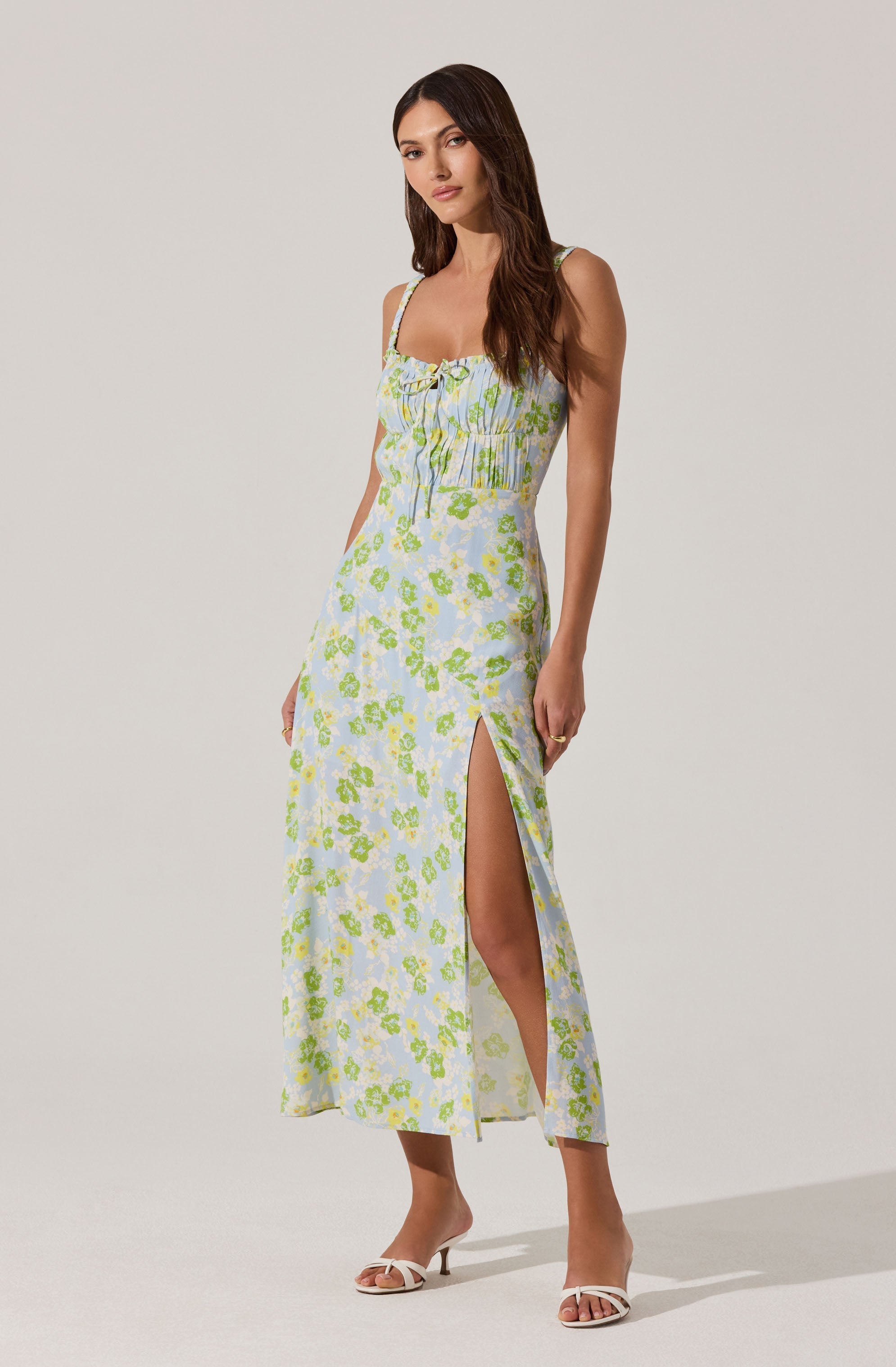 A woman is wearing a sleeveless, light-colored maxi dress with a floral pattern in shades of green and blue. The dress features thin straps, a gathered bodice with a bow at the center, and a high slit on one side. She is also wearing white heeled sandals.