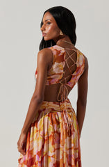 A woman is wearing a sleeveless, floral dress with shades of pink, orange, and cream. The top features a lace-up back with thin ties, creating a crisscross pattern, and the fabric appears lightweight and flowy. The skirt is gathered at the waist, with a wide waistband and a pleated, flowing design. She accessorized with a gold chain necklace and gold earrings.