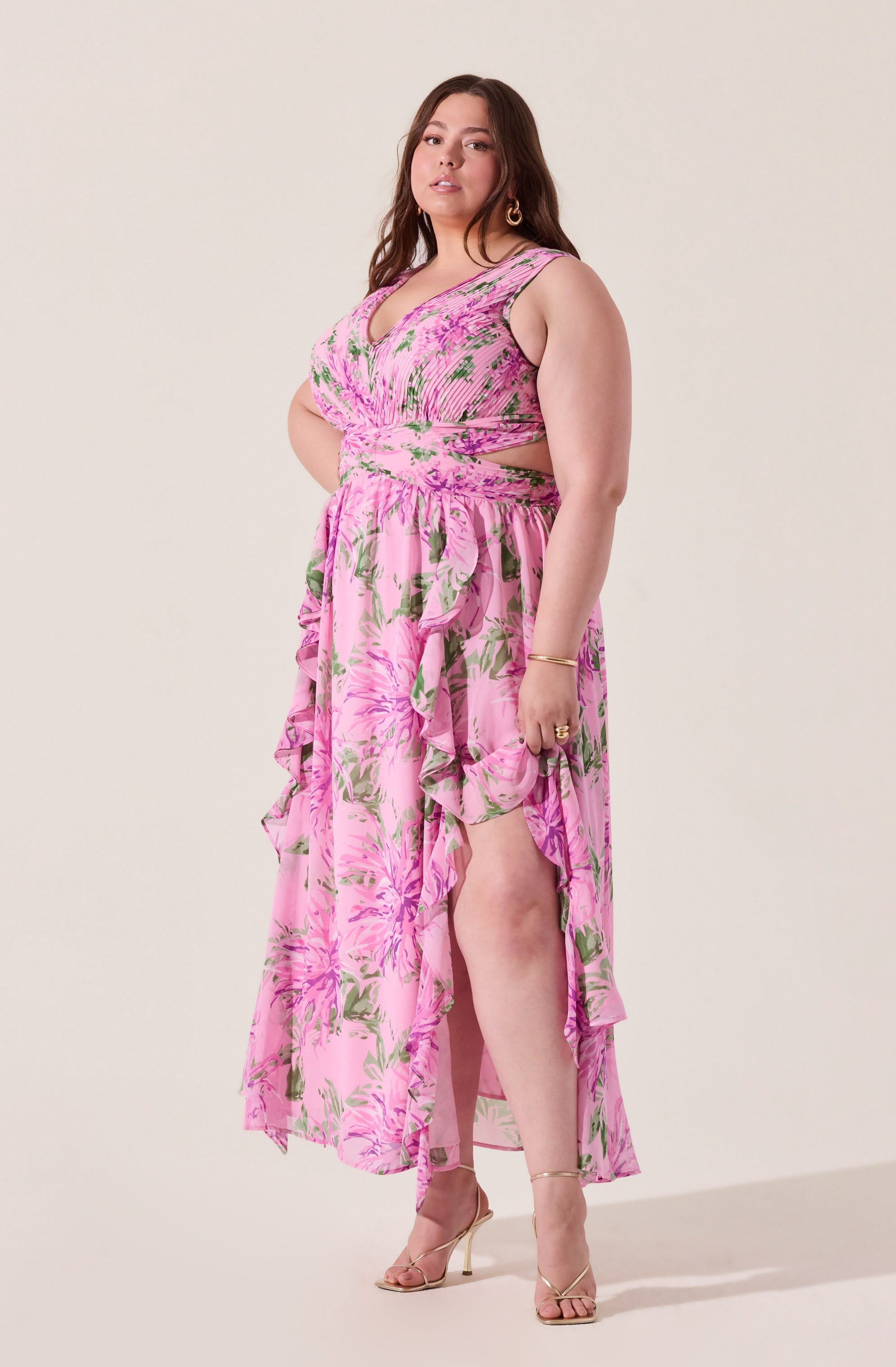 A woman is wearing a pink sleeveless dress with a floral pattern in shades of pink, green, and white. The dress features a fitted bodice with ruching detail and cut-out sides at the waist. The skirt is high-low, with ruffled layers that cascade from the waist to the hem, revealing her legs and gold high-heeled sandals. She accessorizes with gold jewelry, including earrings, a bracelet, and rings.