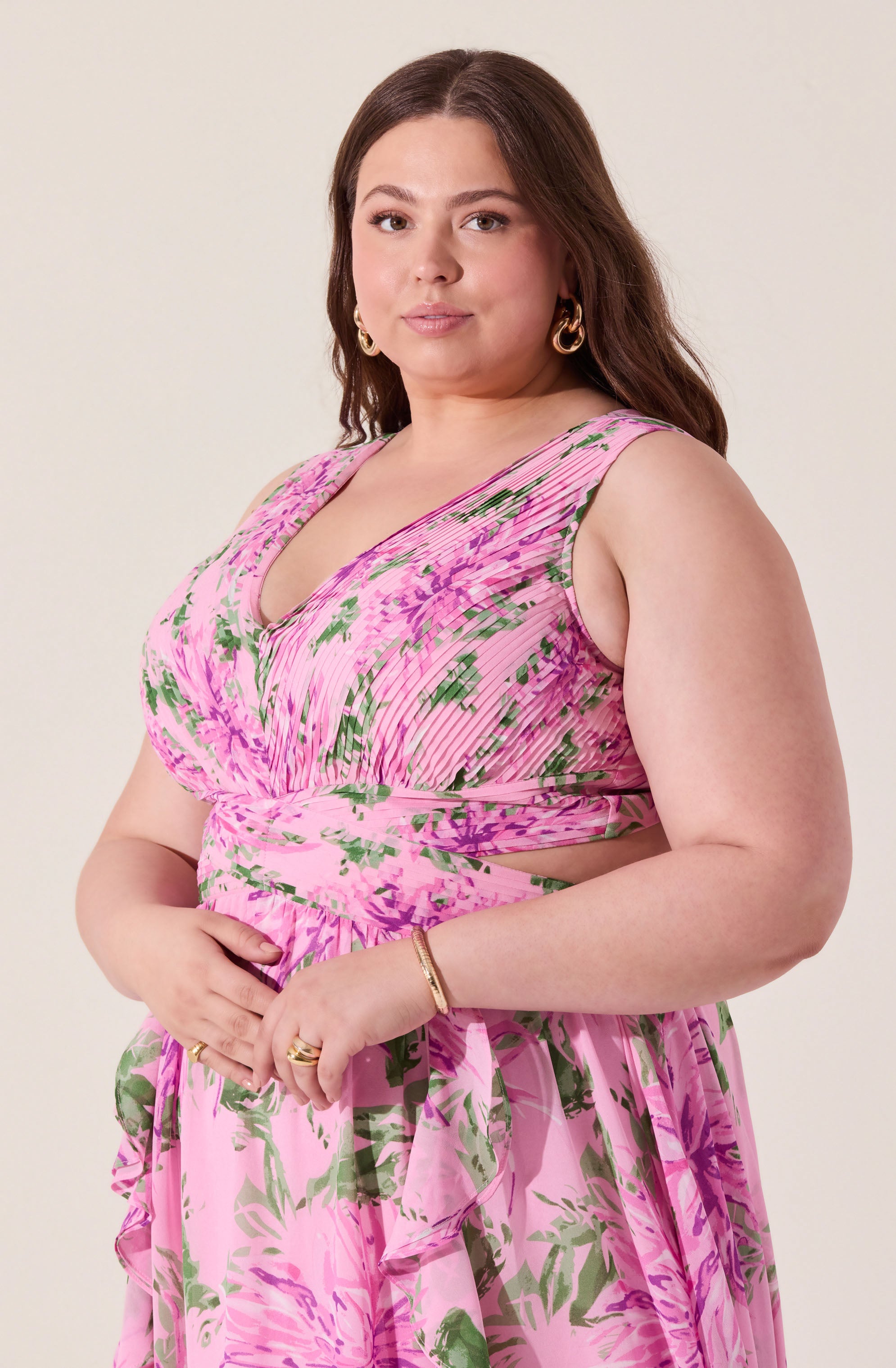A woman is wearing a sleeveless, pink floral dress with green and purple patterns. The dress features a V-neckline, ruching at the bust, and a cutout at the waist.
