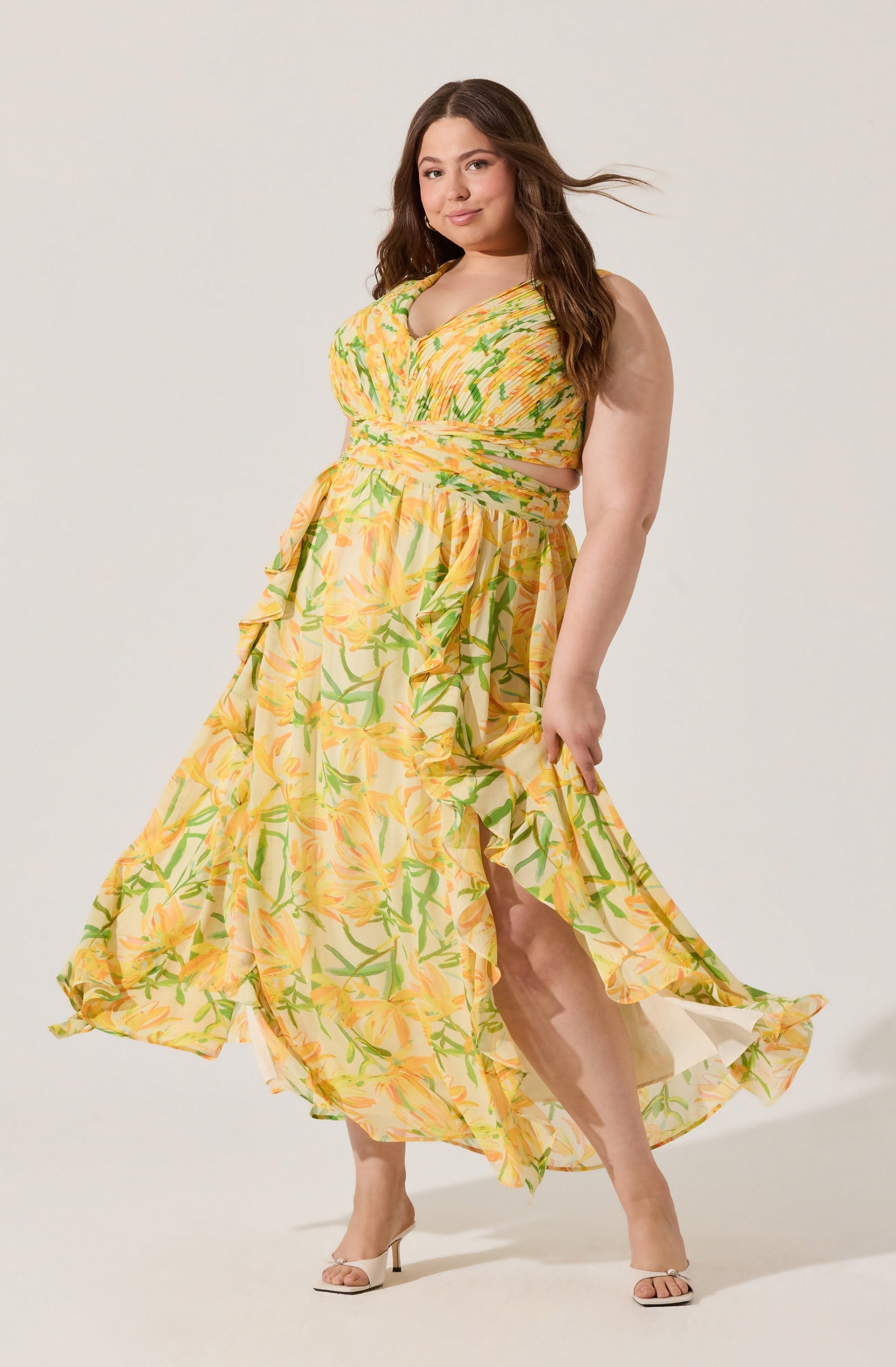A woman is wearing a sleeveless, V-neck maxi dress with a yellow and green floral pattern. The dress features a smocked bodice, a high slit revealing her leg, and a flowing, ruffled skirt. She pairs the outfit with beige high-heeled sandals.