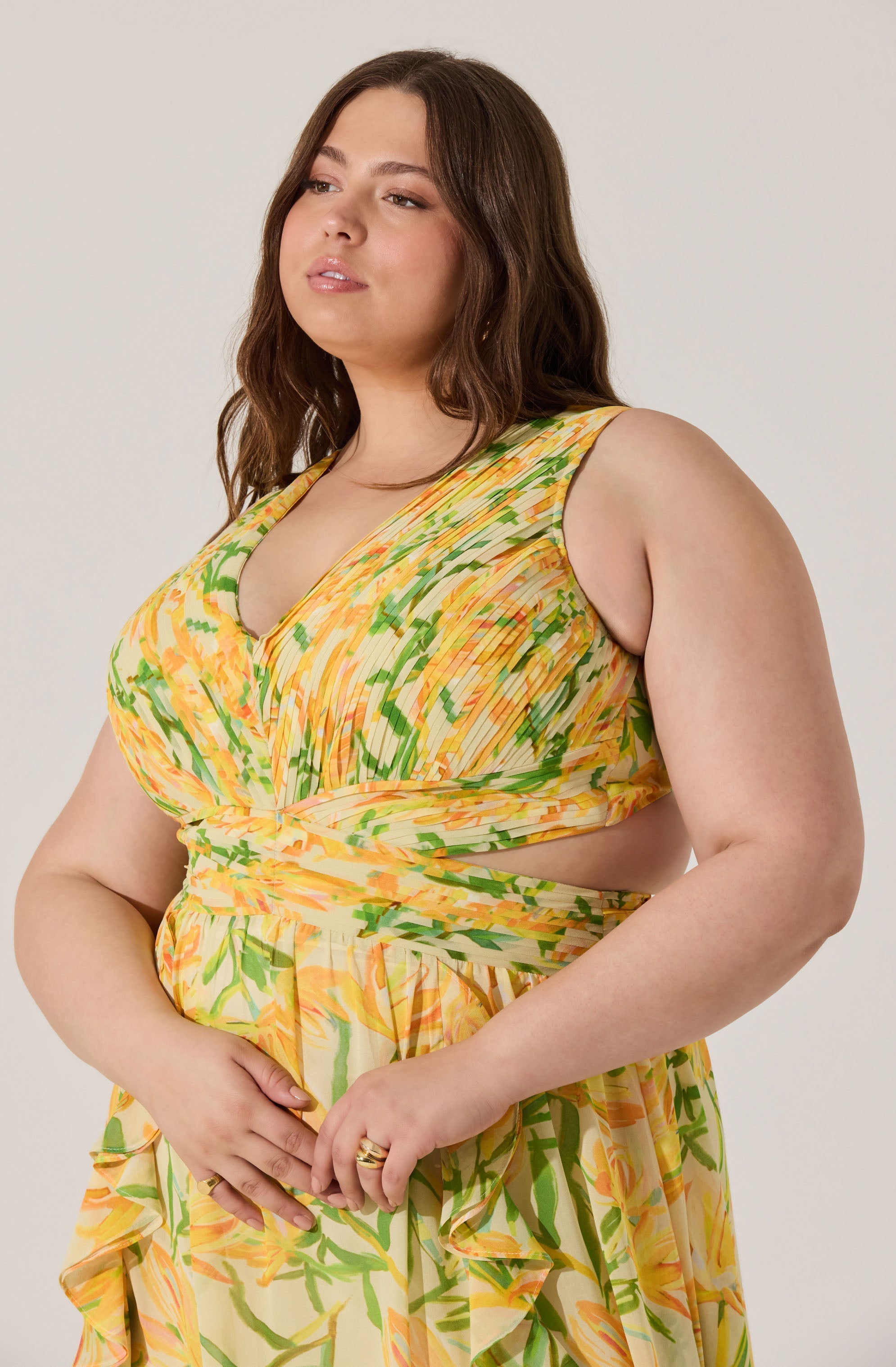 A woman is wearing a sleeveless dress with a vibrant, multicolored abstract pattern featuring shades of yellow, green, and orange. The dress has a V-neckline and a gathered waistband, with the fabric flowing into a loose, tiered skirt.