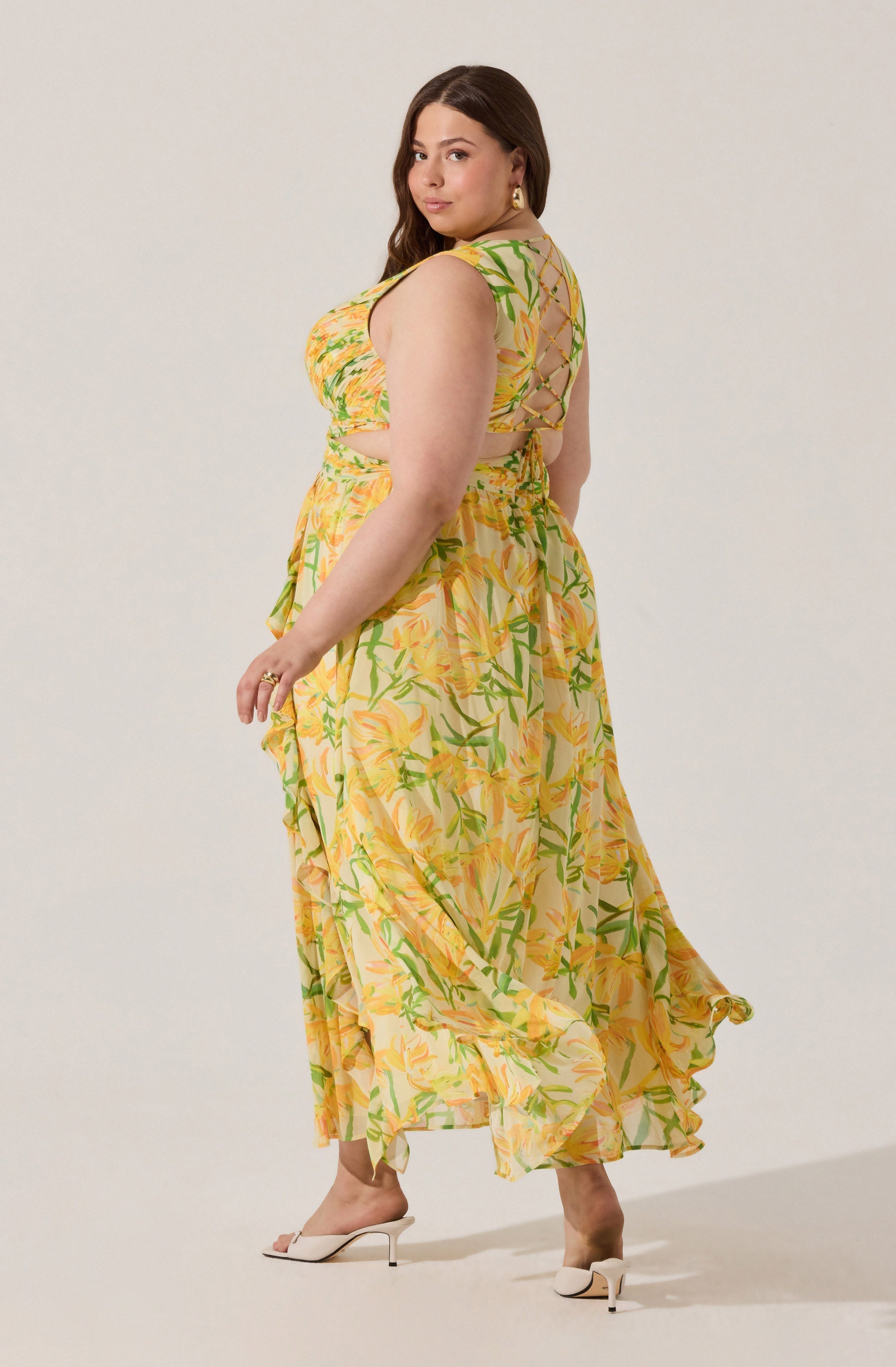 A woman is wearing a sleeveless, flowy maxi dress with a yellow and green floral pattern. The dress features a fitted bodice, a cut-out detail on the back with crisscrossing ties, and a ruffled hem. She is also wearing white high-heeled sandals and gold hoop earrings.