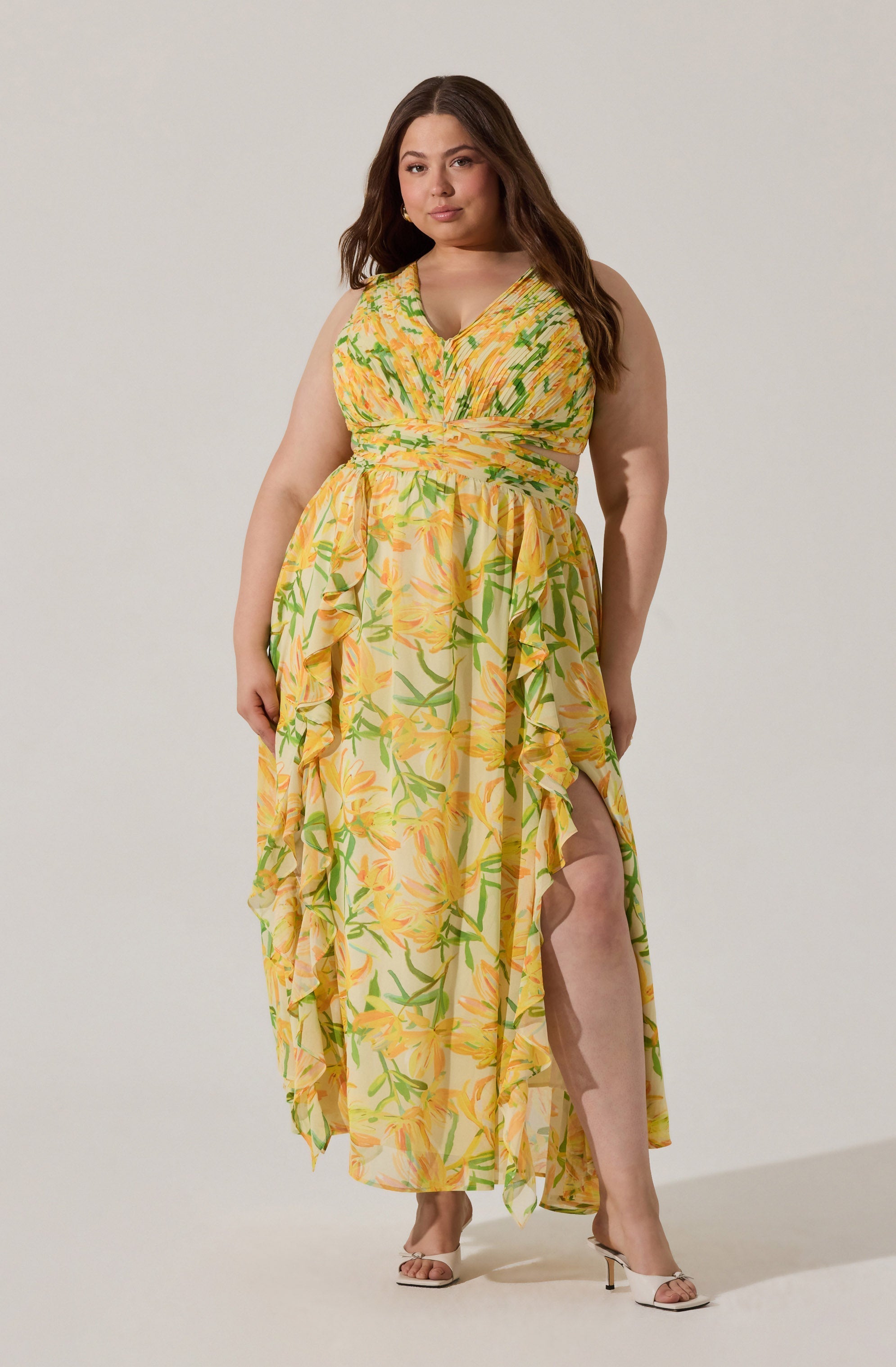 A woman is wearing a sleeveless, V-neck maxi dress with a yellow background and a green and orange floral print. The dress features ruffles along the front slit and the sides, creating a flowing and layered appearance.