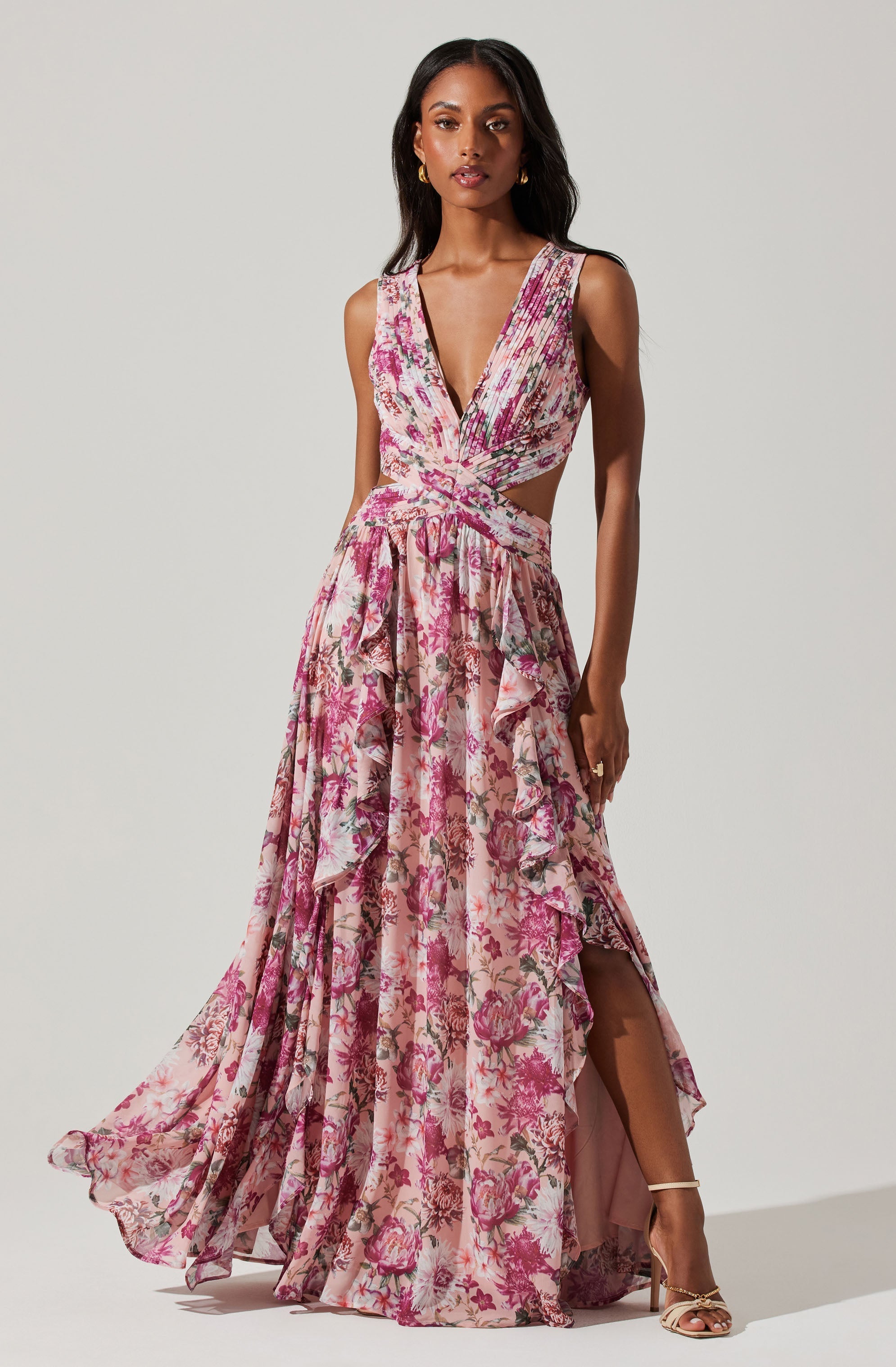 A woman is wearing a sleeveless, deep V-neck gown with a cutout at the waist. The dress features a pink floral pattern on a light background, with ruffled layers and a high slit on one side revealing her leg. She is also wearing gold heeled sandals and gold hoop earrings.
