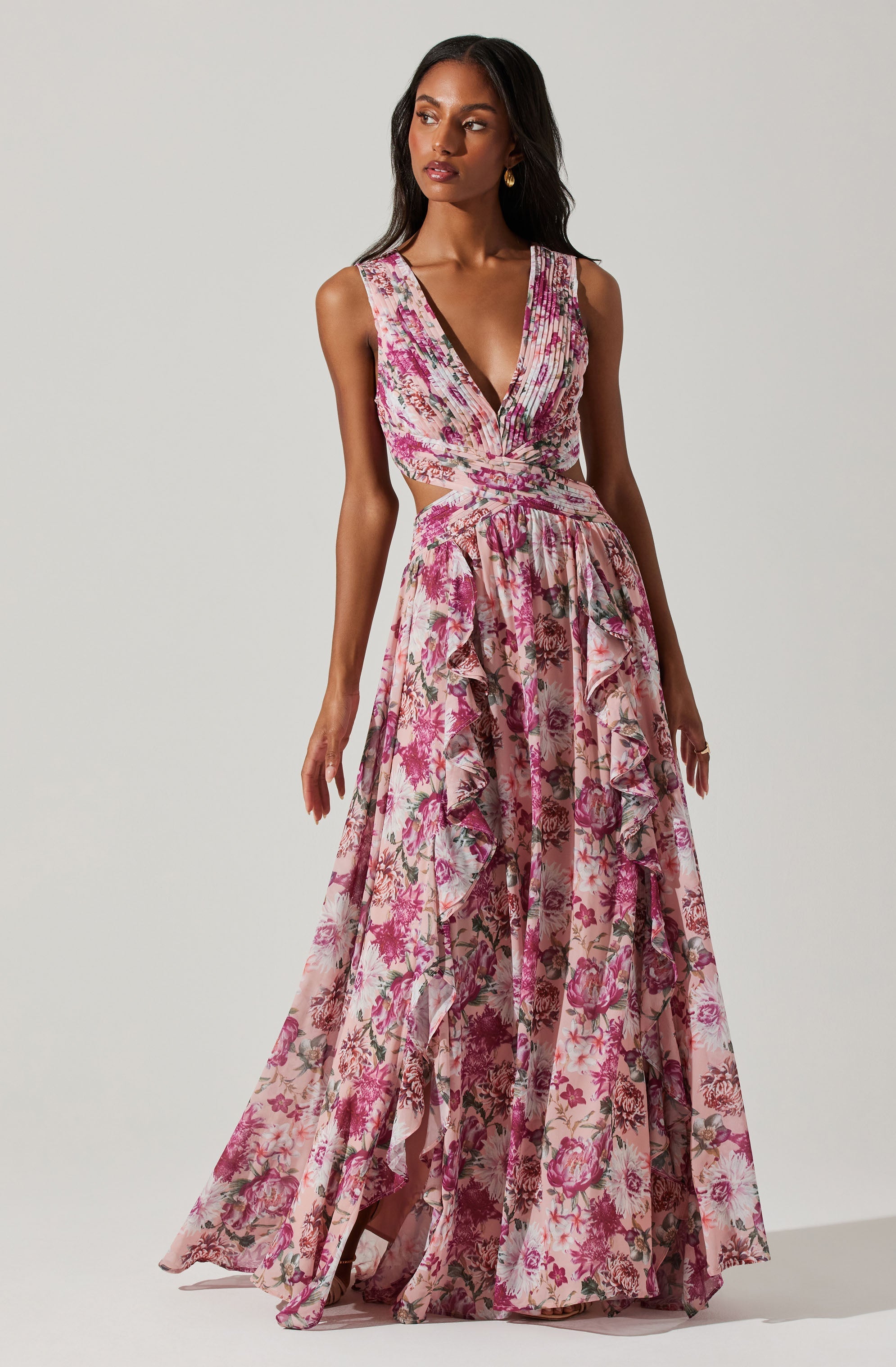 A woman is wearing a sleeveless, V-neck gown with a fitted bodice. The gown features a floral print in shades of pink, purple, and cream. The skirt is long and flowing, with layered ruffles that cascade down the full length of the dress. The fabric appears lightweight and semi-sheer, with a soft, romantic aesthetic.