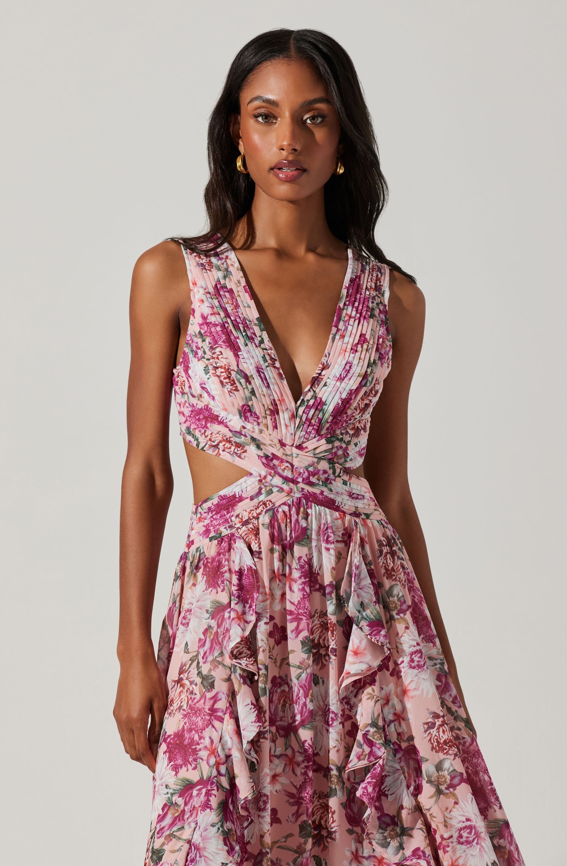 A woman is wearing a sleeveless, V-neck dress with a floral print in shades of pink, purple, and green. The dress features cut-out sides at the waist and has ruffled layers on the skirt.
