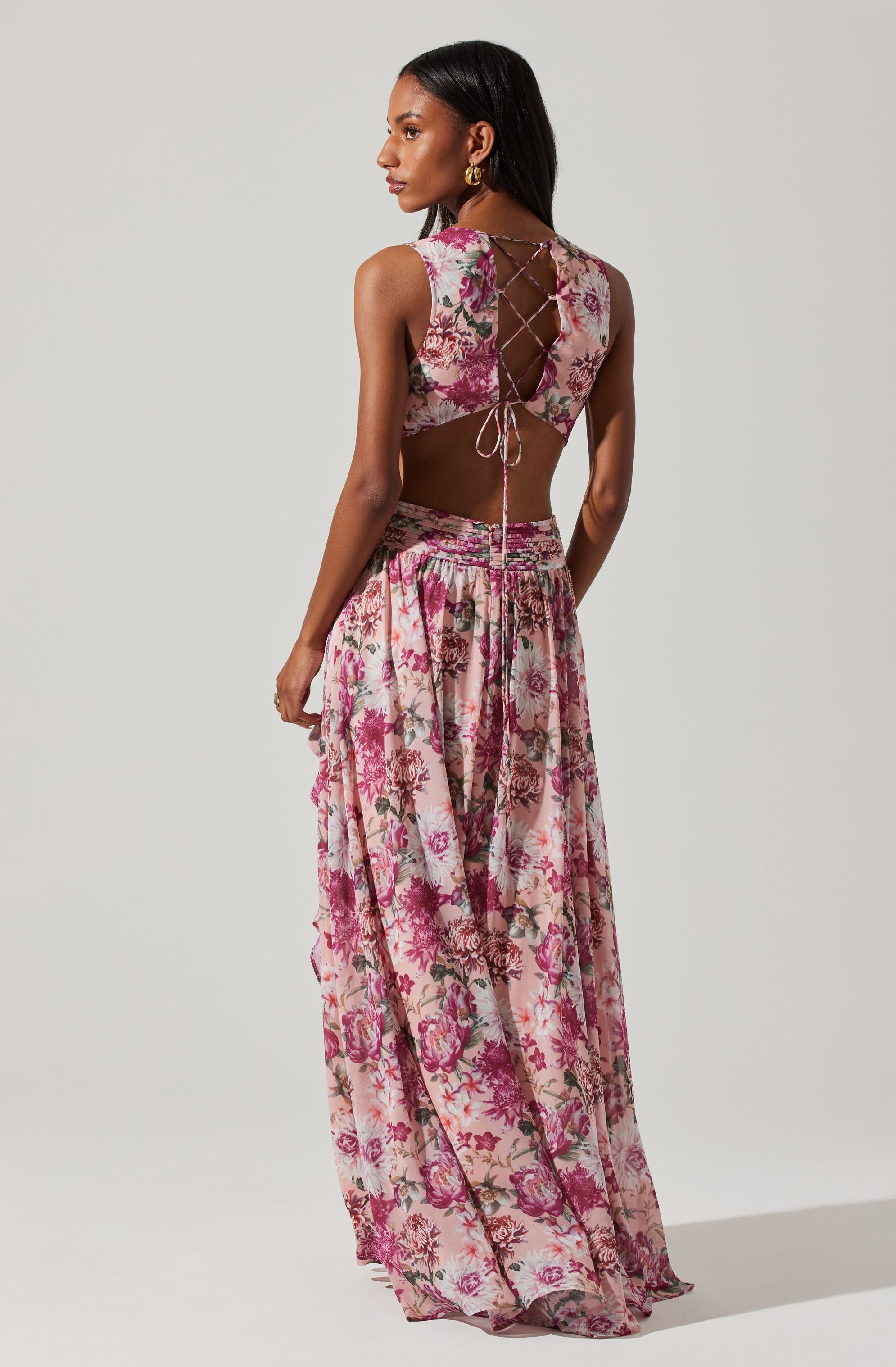 A woman is wearing a sleeveless crop top and a long, flowing skirt, both made from a light fabric with a floral pattern featuring shades of pink, purple, and cream. The crop top has a lace-up back with ties that crisscross and tie at the bottom. The skirt has an elastic waistband and a gathered, relaxed fit that reaches the floor.
