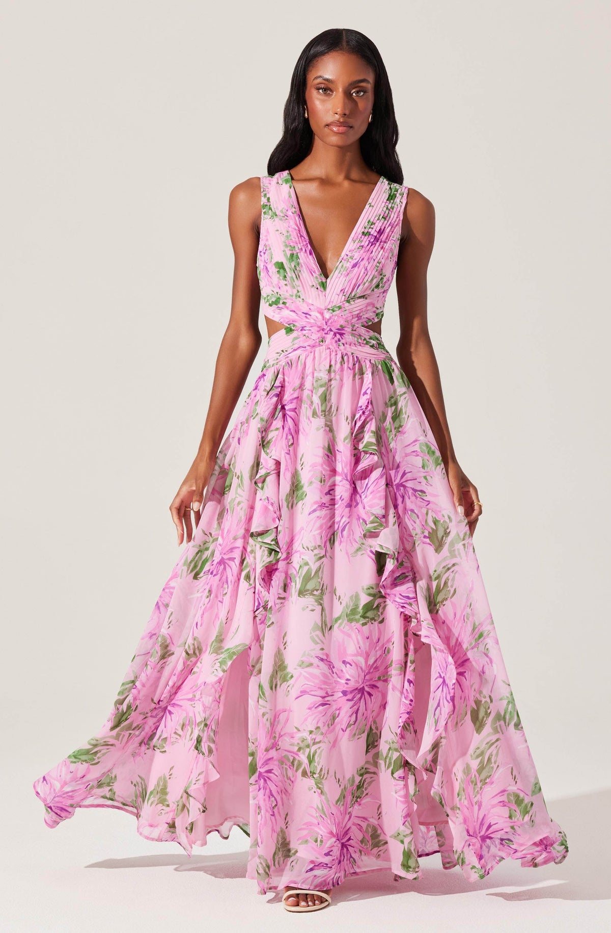 A woman is wearing a sleeveless, V-neck, floor-length dress with a pink floral pattern. The dress has cutouts at the waist and features a flowing, layered skirt with ruffles. The fabric is lightweight and sheer, creating an elegant and airy appearance.