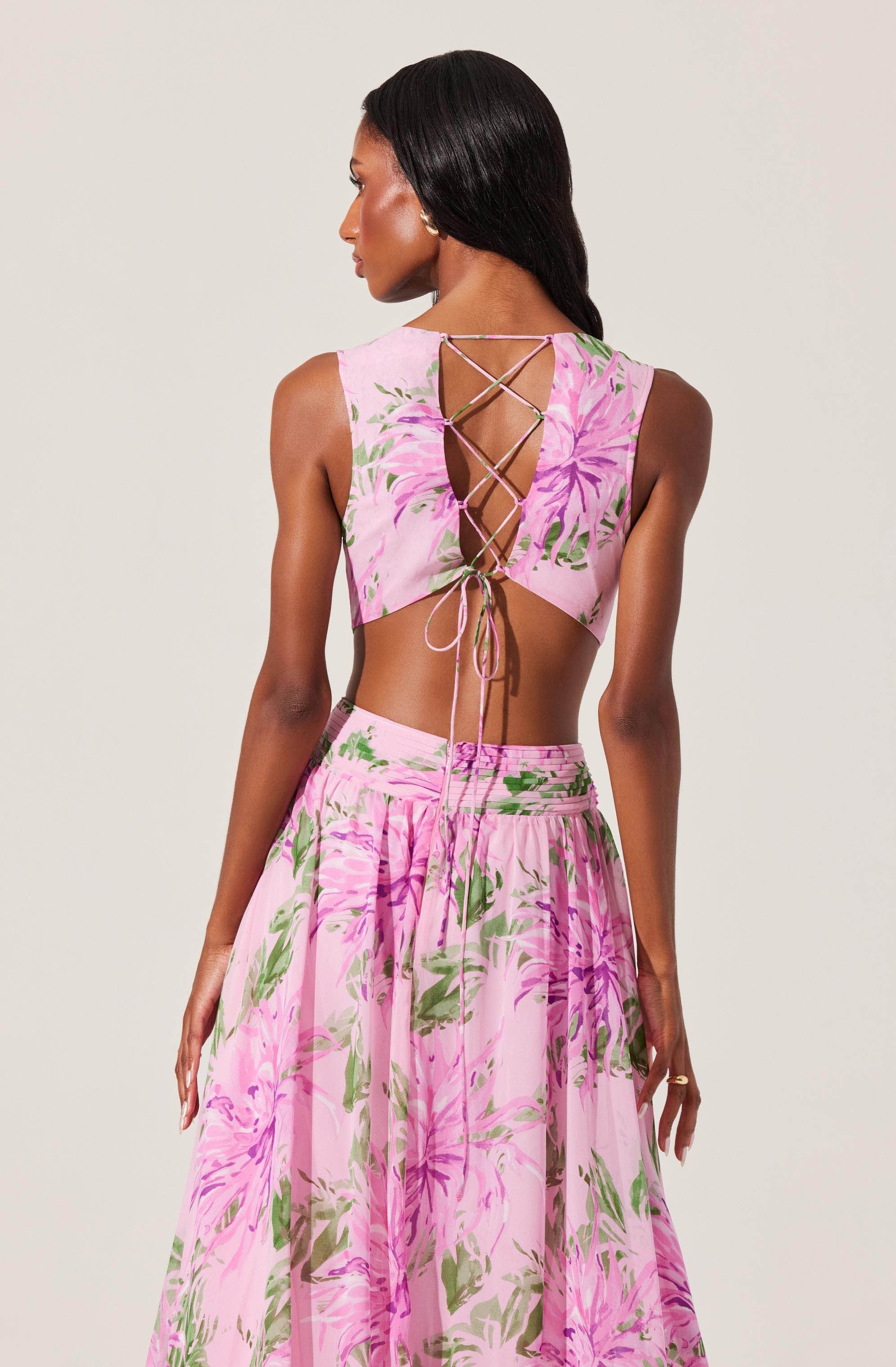 The image features a woman wearing a two-piece outfit with a light pink background and a large floral pattern in shades of pink, purple, and green. The top has a sleeveless design with a cut-out back that is laced up with thin strings crisscrossing down and tied at the bottom. The skirt is high-waisted and flowing, matching the floral pattern of the top.