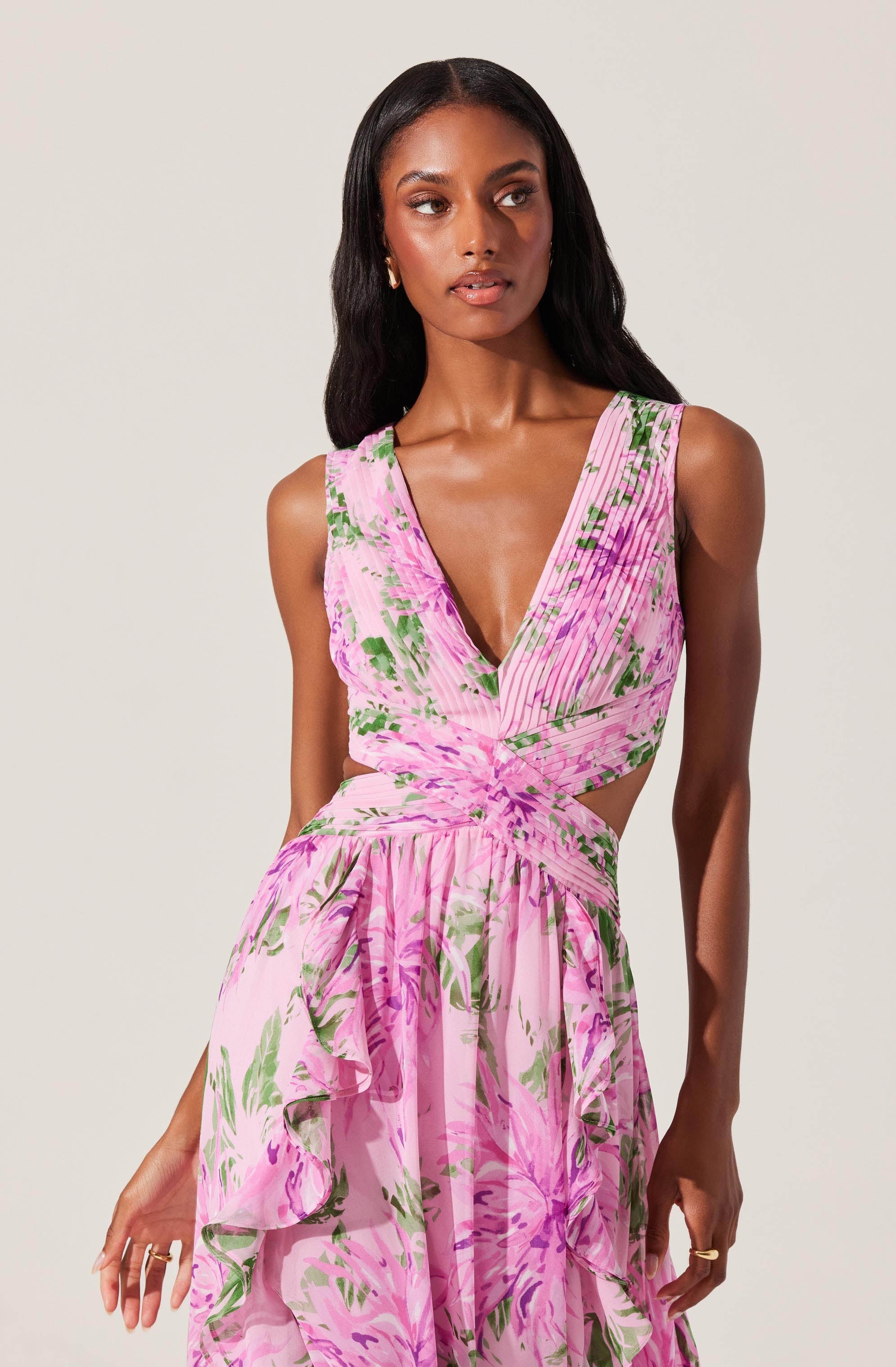 A woman is wearing a sleeveless, V-neck dress with a pink and green floral pattern. The top has a ruched design with cutouts at the waist, and the skirt is flowy with ruffled layers.