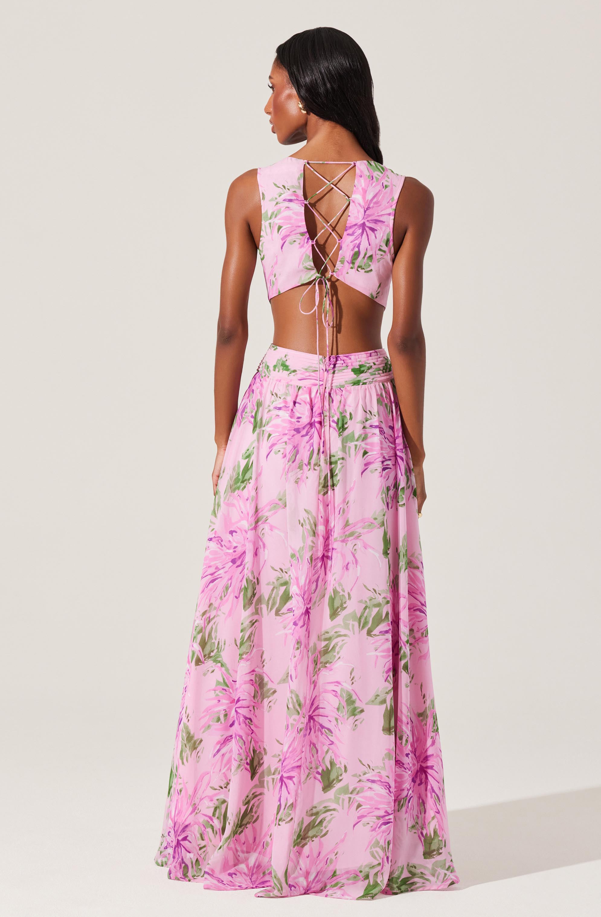 A woman is wearing a two-piece floral outfit consisting of a sleeveless crop top and a long, flowing skirt. The top features a lace-up back with crisscrossing strings, and both garments are decorated with a pink and green floral pattern on a light pink background.