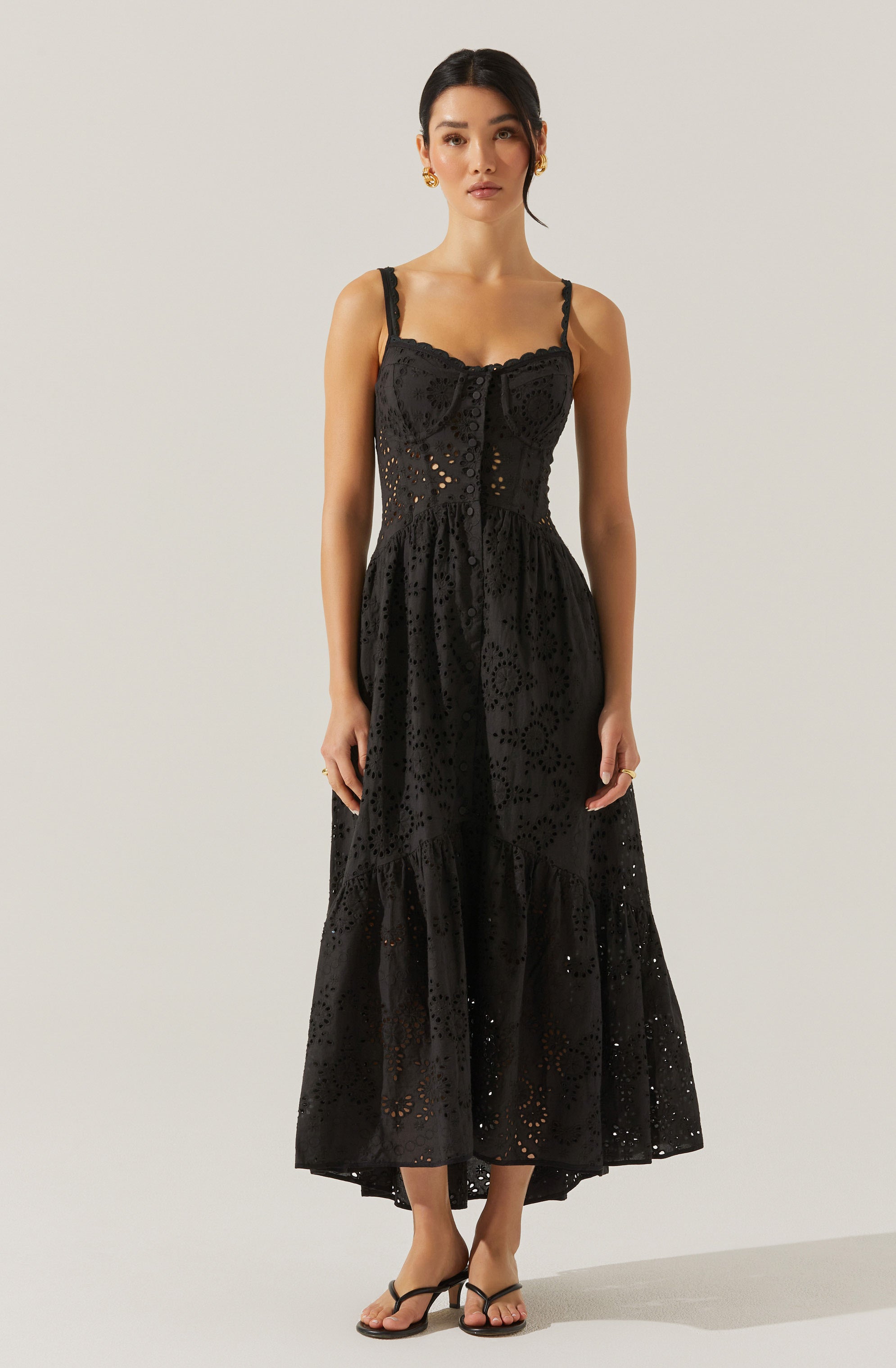 A woman is wearing a black, sleeveless maxi dress with adjustable shoulder straps. The dress features a fitted bodice with a button-down front and an open, eyelet pattern throughout the fabric. The skirt is flowy, gathered at the waist, and has a tiered design with a slightly ruffled hem. She is wearing black sandals with thin straps and small heels.
