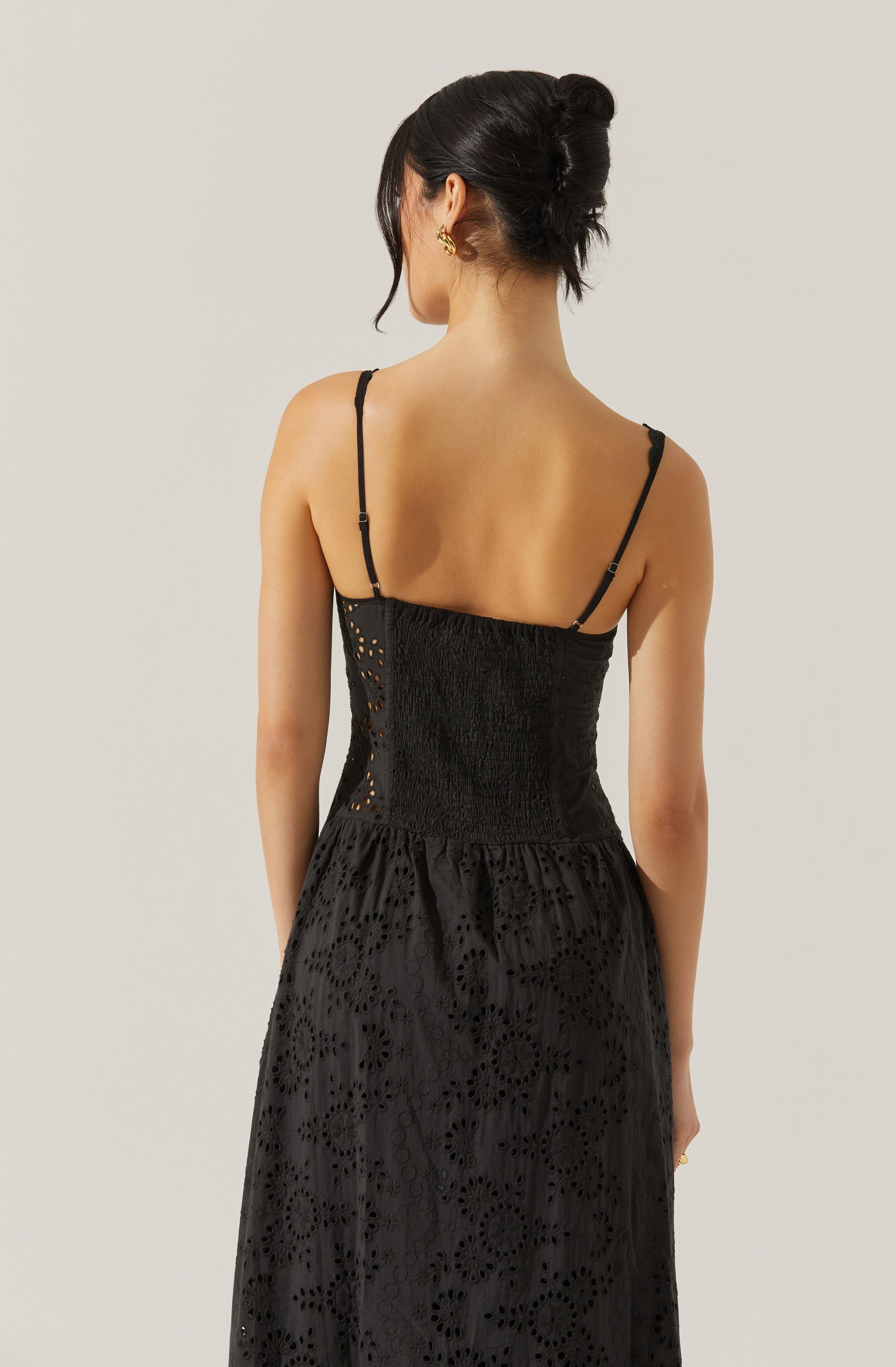 A woman is wearing a black spaghetti strap dress with a fitted, smocked bodice that features a broderie anglaise pattern with eyelet detailing. The skirt portion of the dress is flowy and also adorned with eyelet patterns, creating a textured, intricate look. The dress has thin, adjustable straps that rest on her shoulders.