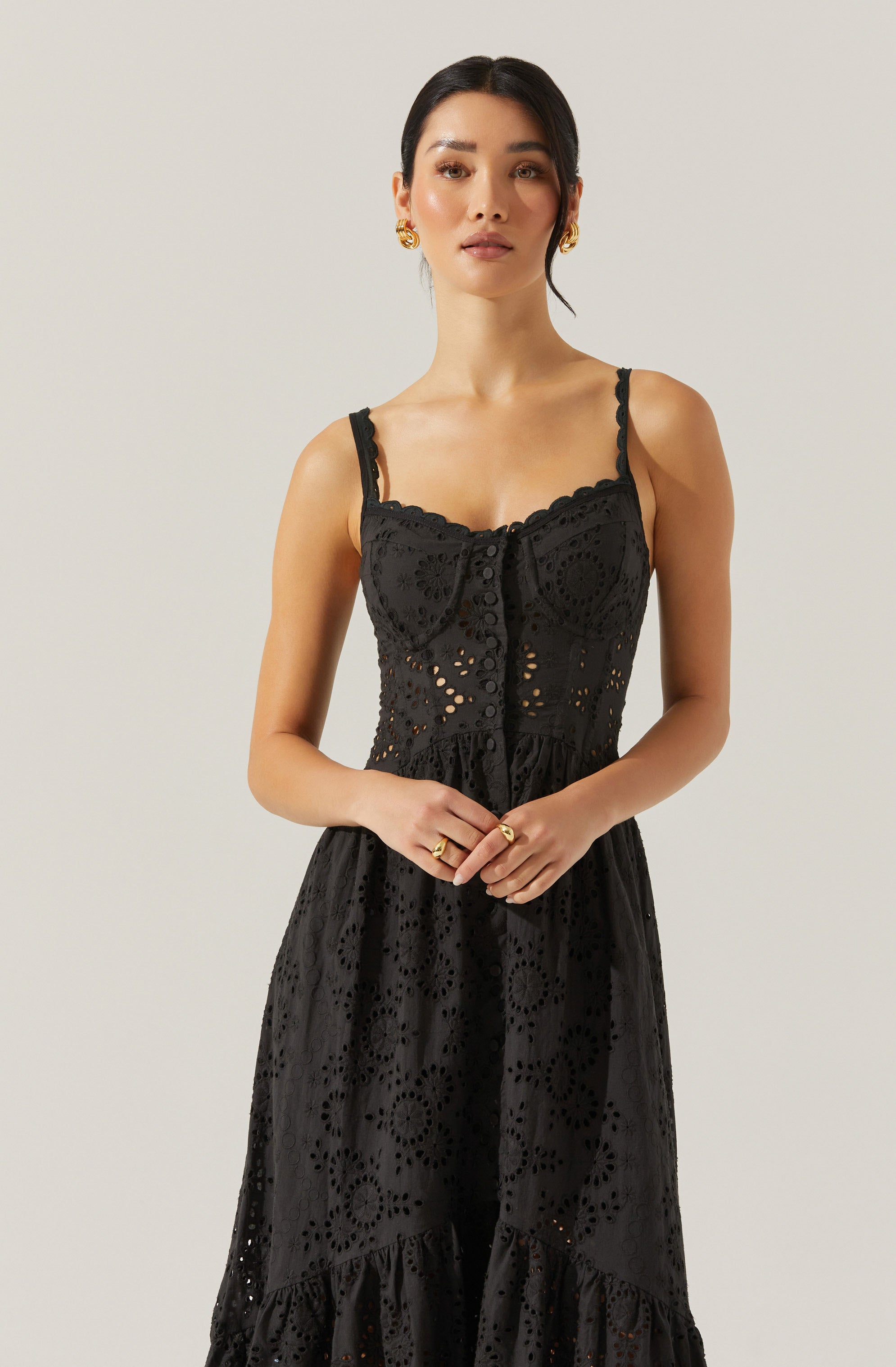 A woman is wearing a black sleeveless dress with spaghetti straps. The dress features intricate eyelet embroidery with floral and circular patterns throughout, a fitted bodice with buttons down the front, and a ruffled detail at the neckline. The skirt is long and flowing, with a ruffle at the hem. She is accessorized with gold earrings and rings.