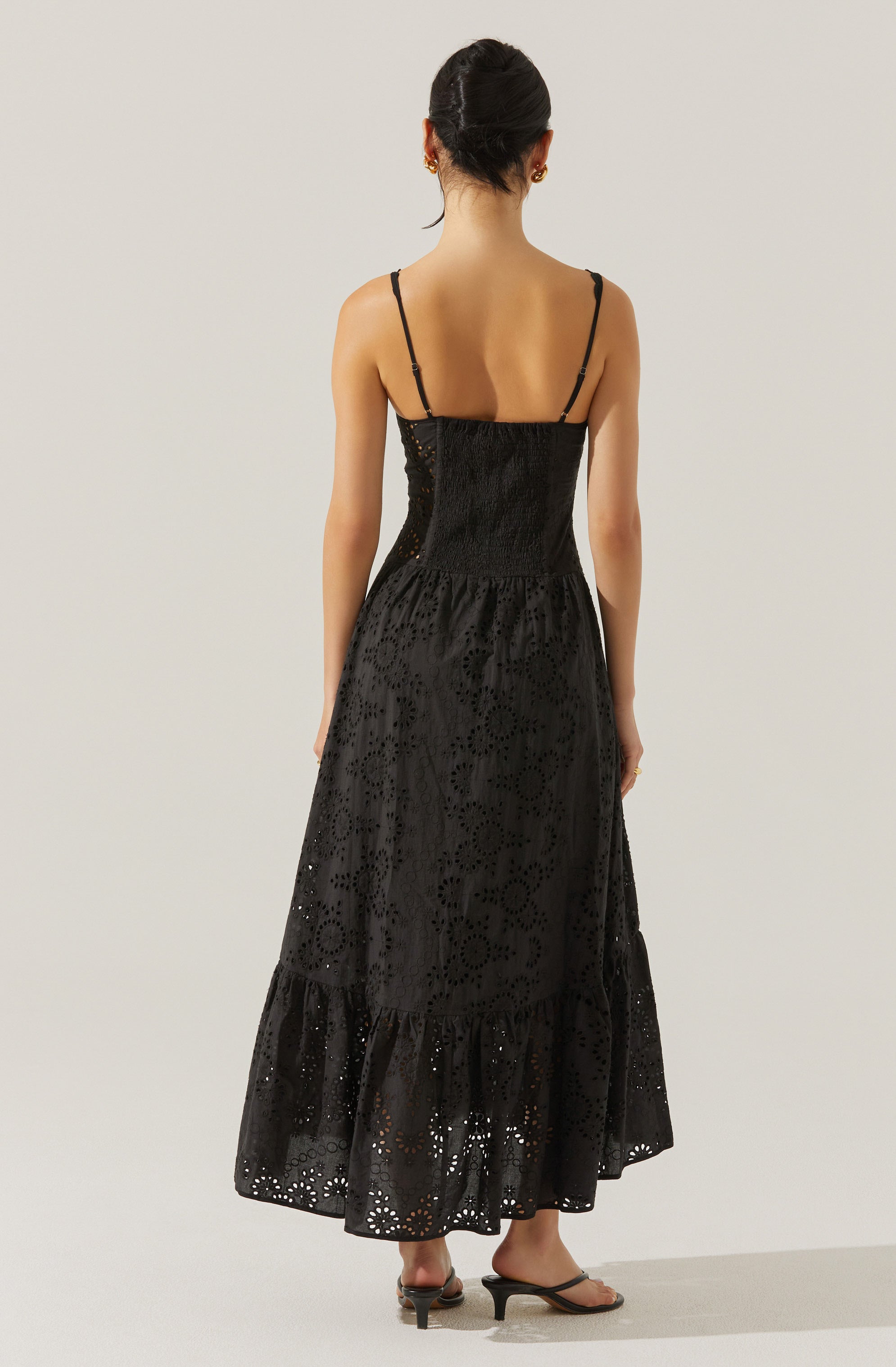 A woman is wearing a long, black, sleeveless dress with thin spaghetti straps. The dress features a fitted bodice and a full-length skirt with eyelet embroidery, creating a textured, patterned appearance. The skirt has a tiered ruffle at the hem, adding a decorative touch.