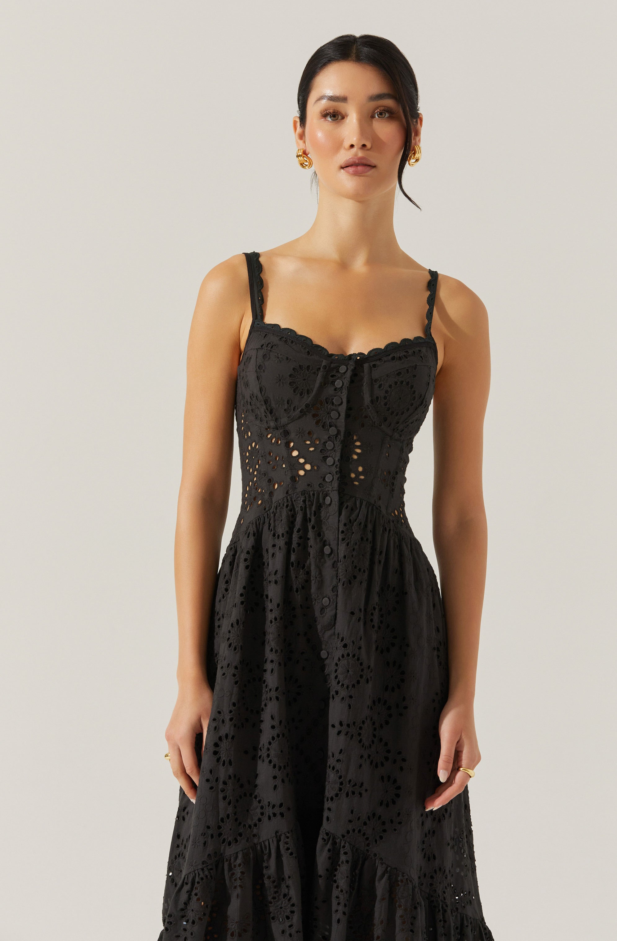 A woman is wearing a black eyelet lace dress with thin, adjustable straps. The dress features a fitted bodice with scalloped edges and buttons down the front, and the skirt has a flowing, tiered design with intricate circular cut-out patterns throughout.