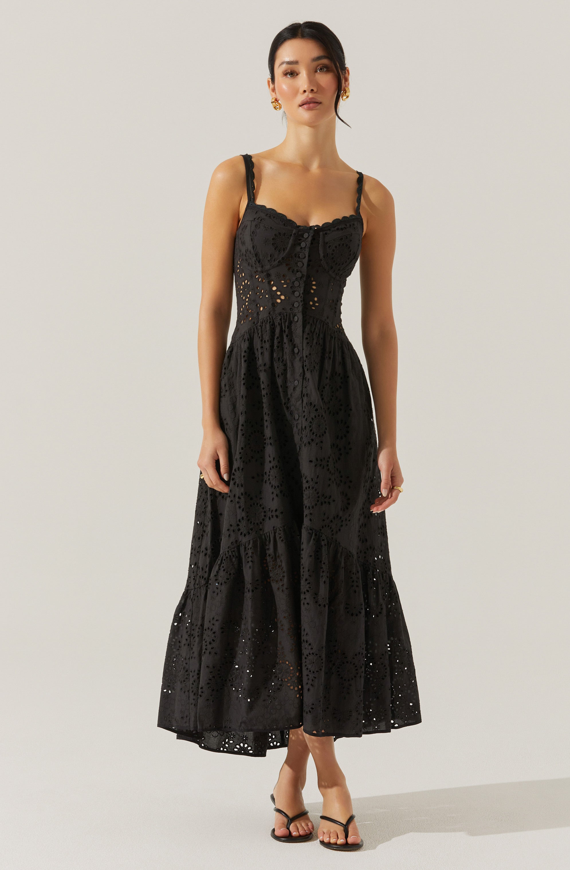 A woman is wearing a black sleeveless dress with thin straps. The dress has a fitted bodice with a ruffled detail at the neckline and features intricate eyelet embroidery throughout. The skirt is long, flowing, and tiered, reaching down to her ankles, with the same eyelet embroidery pattern.