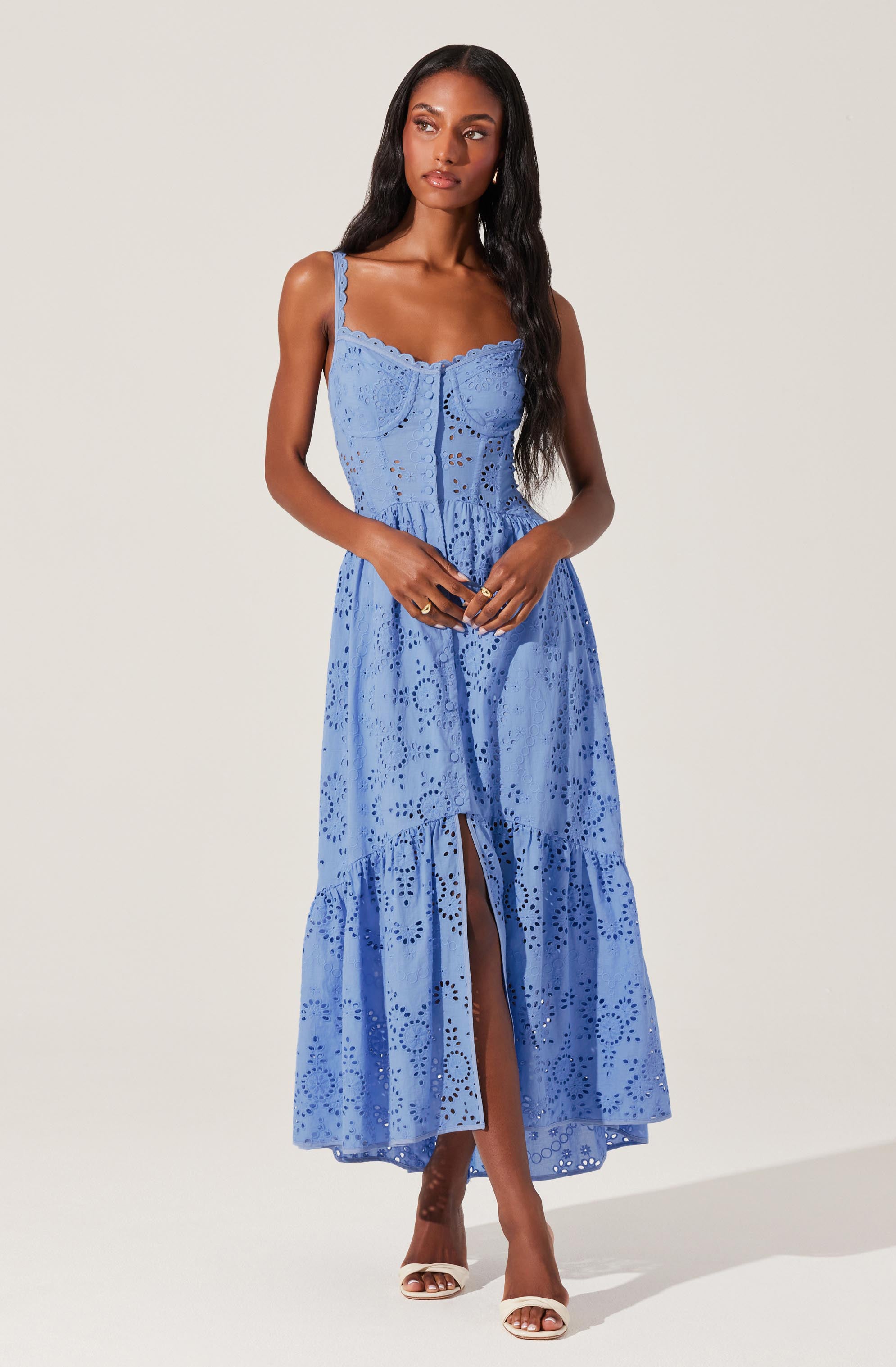 A woman is wearing a sleeveless, light blue dress with adjustable straps. The dress features intricate eyelet embroidery throughout, creating a pattern of floral and geometric shapes. It has a fitted bodice with a sweetheart neckline and a button detail down the front. The skirt is long, flowing, and tiered, with a slit up the front revealing her legs. She is wearing white high-heeled sandals.