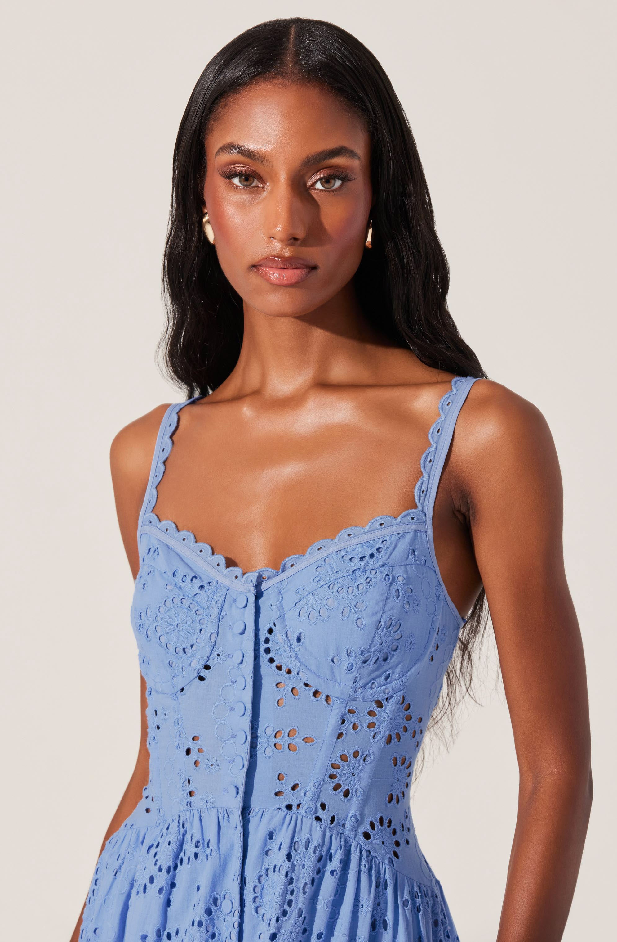 A woman is wearing a sleeveless, light blue dress with embroidered eyelet details. The dress features scalloped edges along the straps and neckline, with intricate cut-out patterns throughout the fabric.