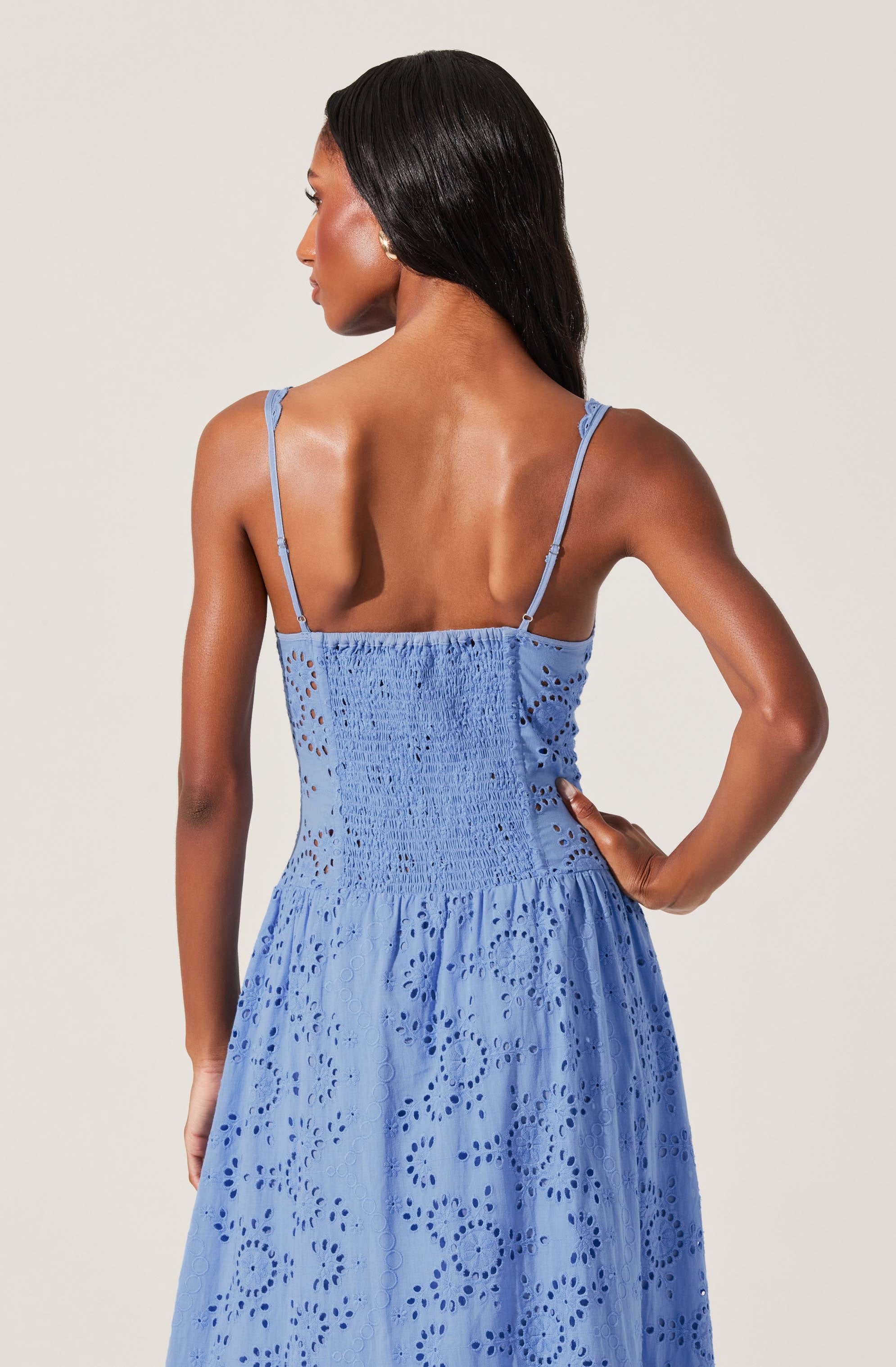 A woman is wearing a light blue dress with thin spaghetti straps. The dress features intricate eyelet embroidery throughout, creating a pattern of circular and floral designs. The bodice is smocked for a fitted look, and the skirt flows loosely. The fabric appears to be lightweight and suitable for warm weather.