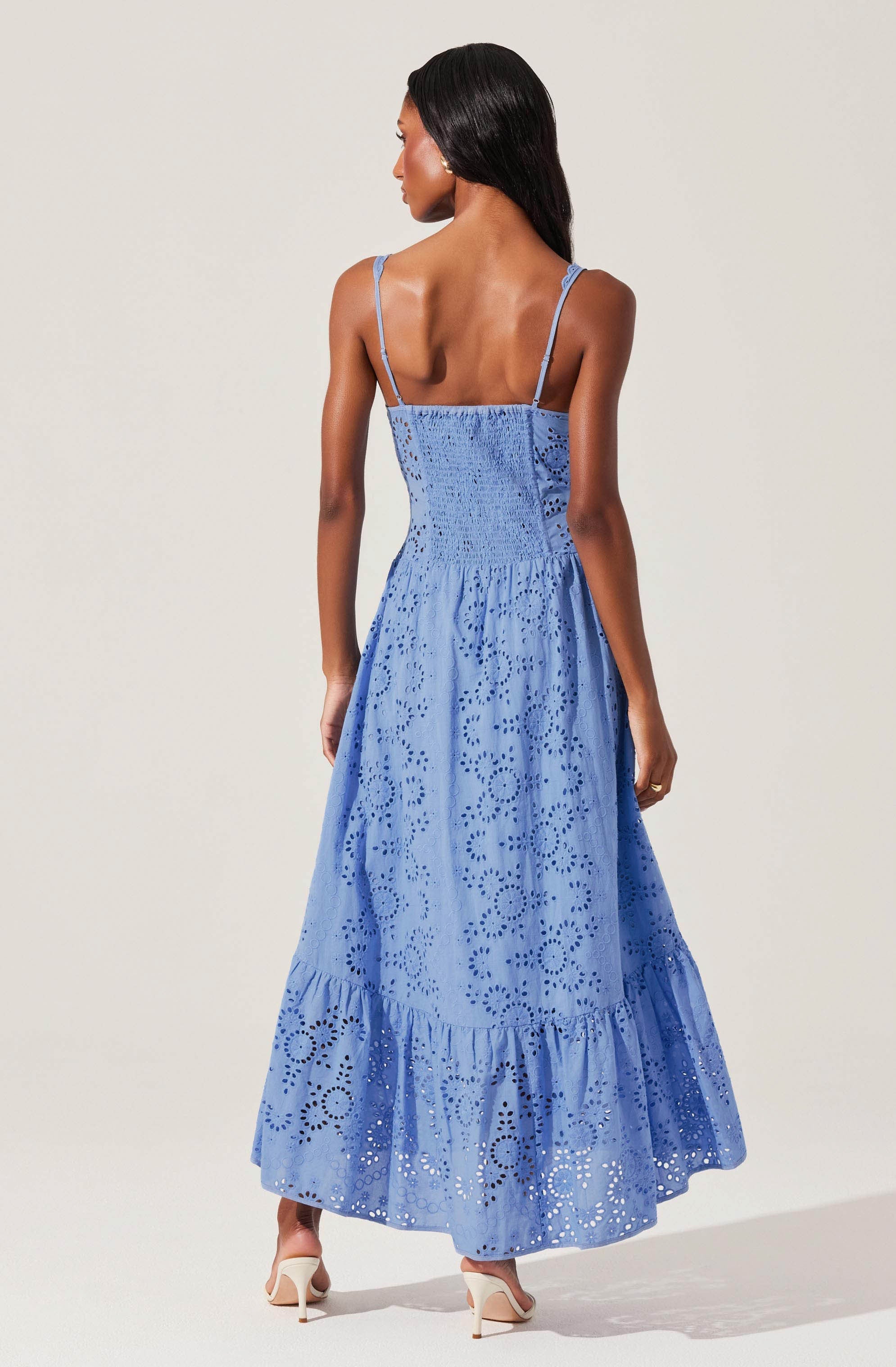 A woman is wearing a sleeveless, ankle-length blue dress with spaghetti straps. The dress features an eyelet pattern with circular and floral cutouts throughout the fabric, creating a textured, decorative appearance. The waistline is fitted, and the skirt is gathered, adding volume and a tiered ruffle detail at the hem.