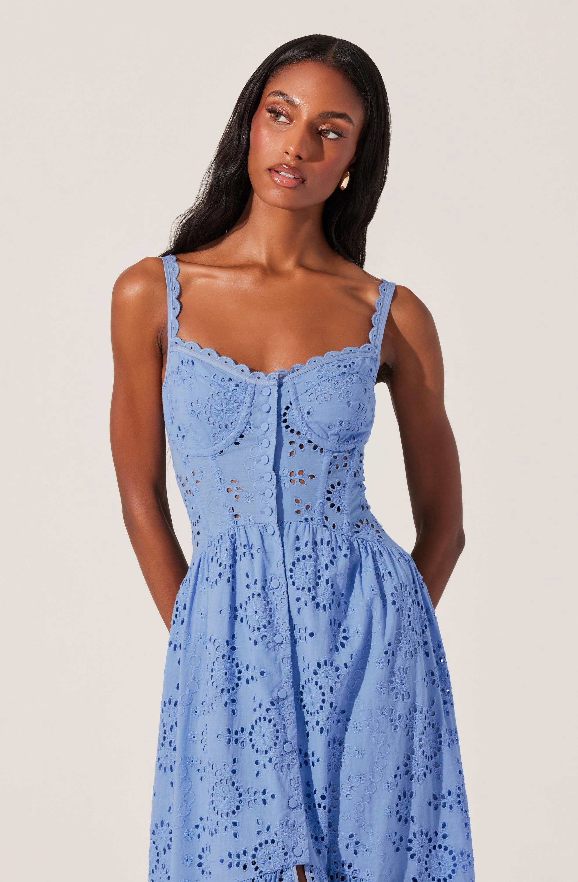 A woman is wearing a sleeveless, blue dress with intricate eyelet embroidery. The dress has adjustable straps, a fitted bodice with decorative buttons, and a gathered skirt that falls below the waist. The eyelet pattern features circular and floral designs throughout the fabric.