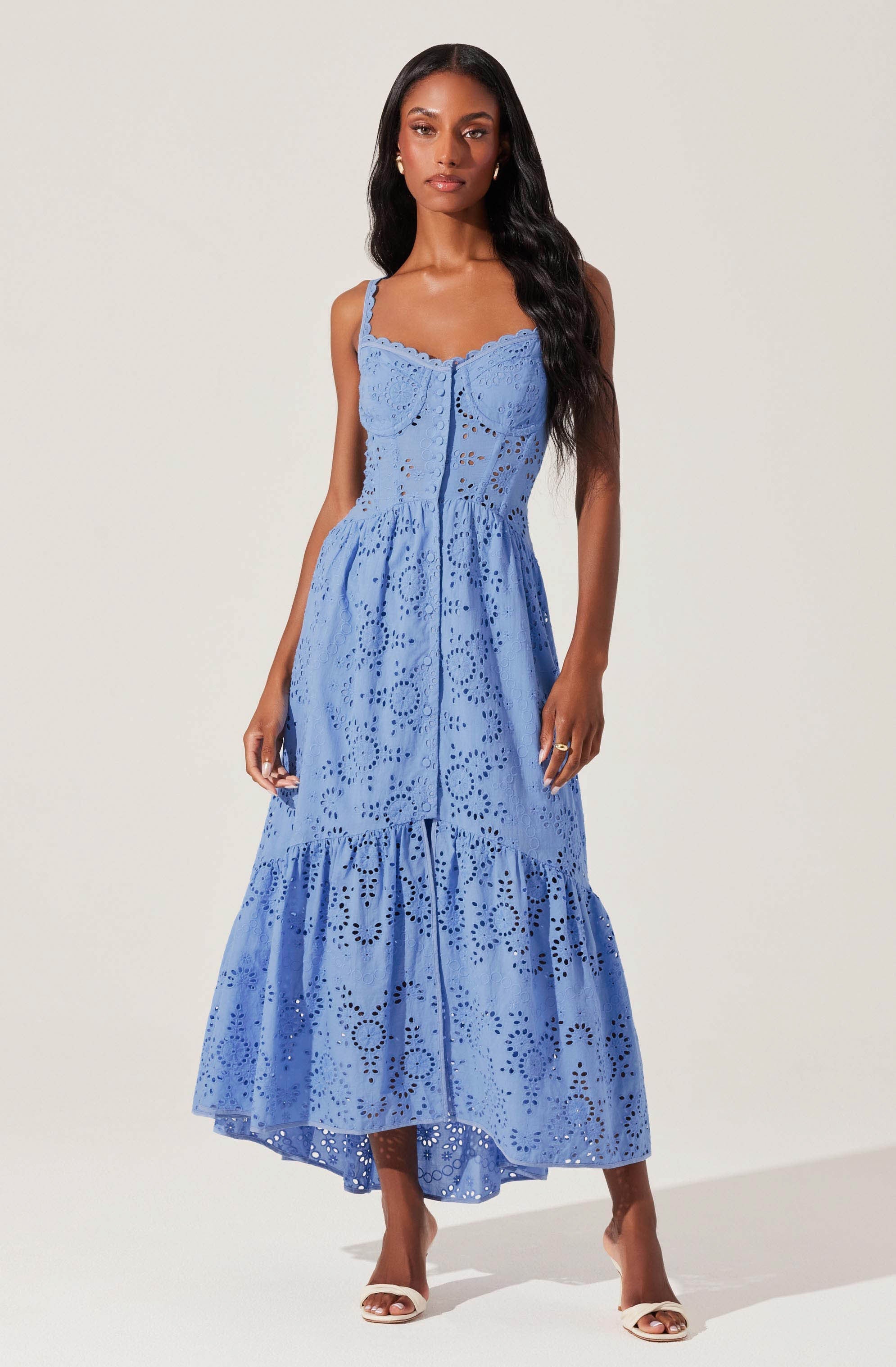A woman is wearing a long, sleeveless, light blue dress made of eyelet fabric with cut-out patterns. The dress features a fitted bodice with a sweetheart neckline and adjustable straps, and it has a tiered, A-line skirt that extends to her ankles. She is standing against a plain light background, accessorized with earrings and white open-toe heels.