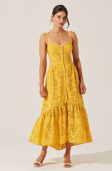 A woman is wearing a yellow, sleeveless, eyelet lace dress with a fitted bodice featuring a front button closure. The dress has thin shoulder straps with scalloped edges and a tiered, flared skirt that reaches just above the ankles. She is also wearing open-toed white high-heeled sandals.