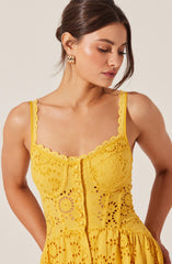 A woman is wearing a sleeveless yellow dress with scalloped edges on the straps and neckline. The dress features embroidered eyelet patterns throughout, with a fitted bodice and a gathered skirt. The front of the dress has a vertical row of buttons running down the center.