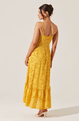 A woman is wearing a bright yellow, sleeveless summer dress with thin spaghetti straps. The dress features eyelet embroidery with circular and floral patterns throughout, and a ruffled hem at the bottom. She is wearing light-colored high heels.
