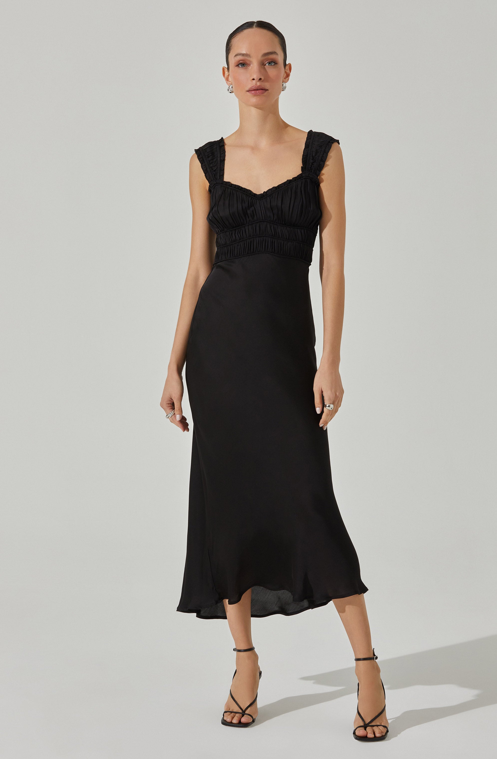 A woman is wearing a long, black, sleeveless dress with a fitted bodice that features ruching and textured fabric on the straps and upper part. The dress has a simple, elegant silhouette and a slightly flared hem. She is accessorizing with silver earrings and rings, and is wearing black high-heeled sandals with ankle straps.