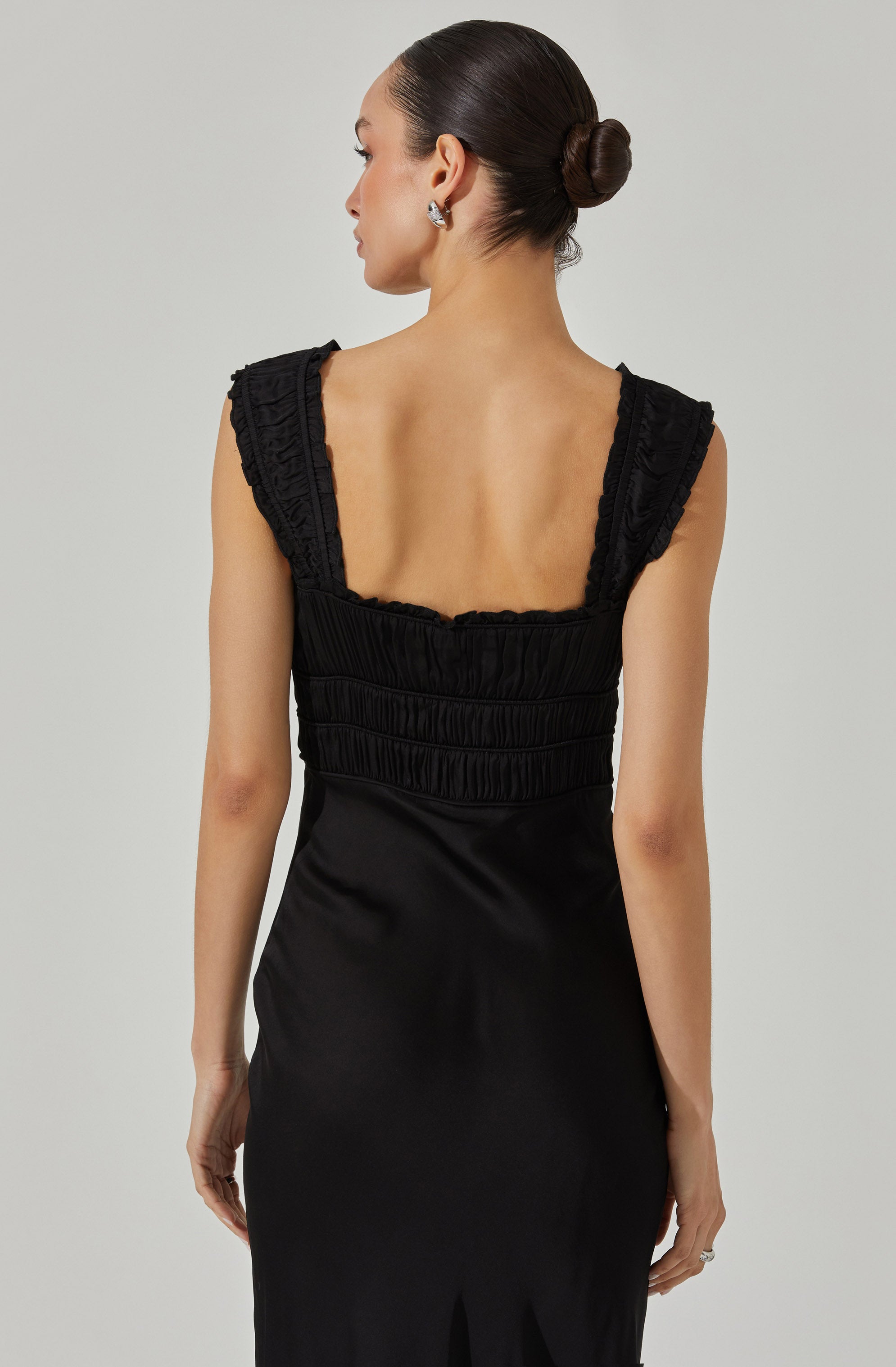 A woman wearing a sleek black dress with ruched detailing on the bodice and ruffled shoulder straps. The dress has a fitted silhouette with a subtle slit at the hem.