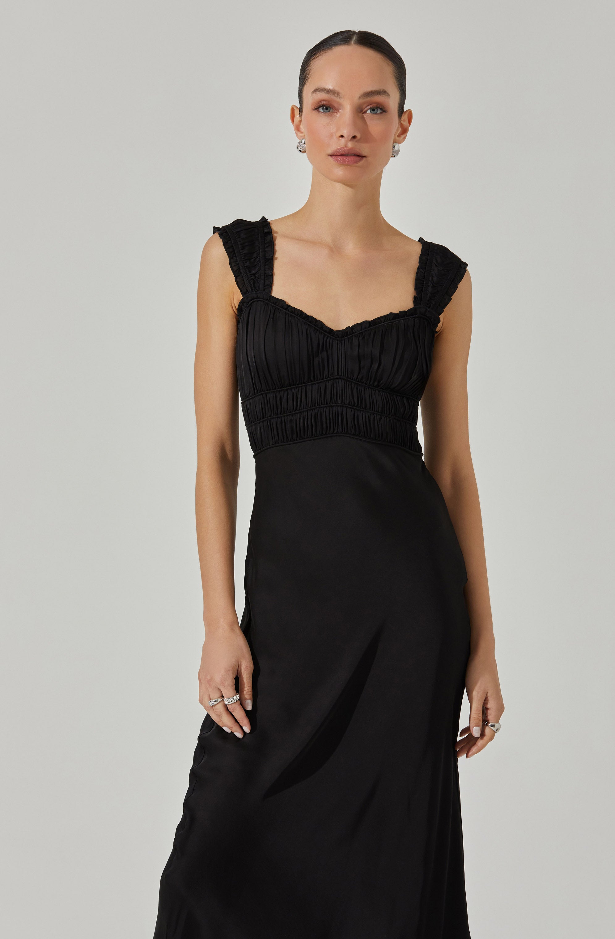 A woman is wearing a sleeveless black dress with ruffled shoulder straps and a ruched bodice. The dress has a fitted waist and a long, flowing skirt. She is also wearing earrings and multiple rings on her fingers.