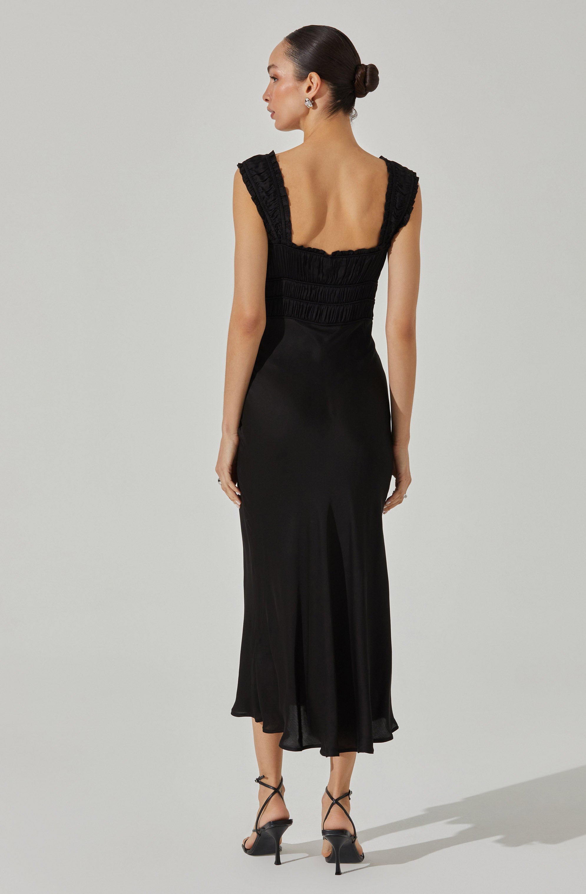 A woman is wearing a black sleeveless dress with textured, ruffled shoulders and a fitted bodice with horizontal ruching. The skirt of the dress is flowy and reaches mid-calf, with a slit at the back. She has her hair styled in a low bun and is wearing earrings. The dress has a sleek and elegant appearance.