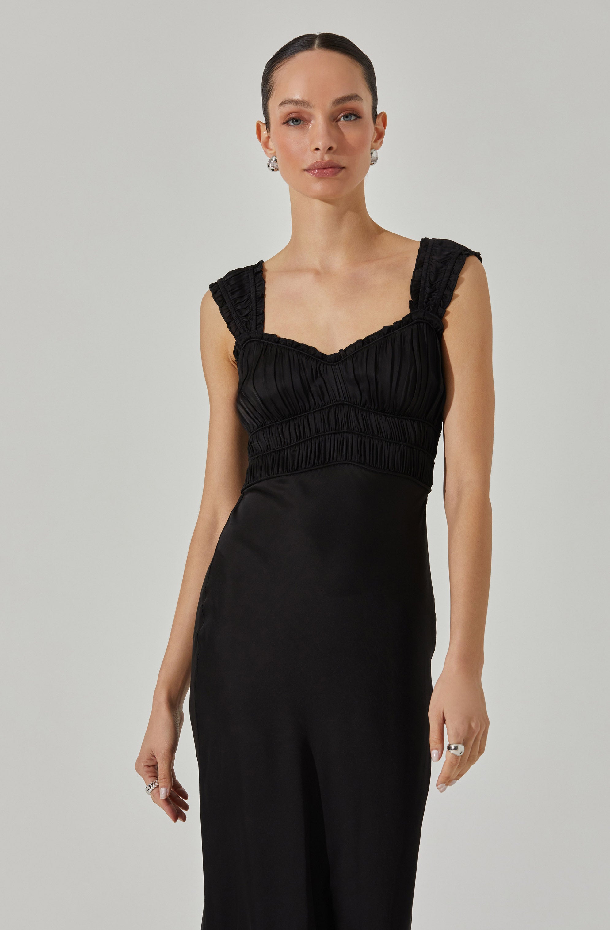 A woman is wearing a sleeveless black dress with ruffled shoulder straps and a fitted bodice with gathered fabric details. The dress has a smooth, flowing skirt. She accessorizes with statement earrings and a ring on her right hand.
