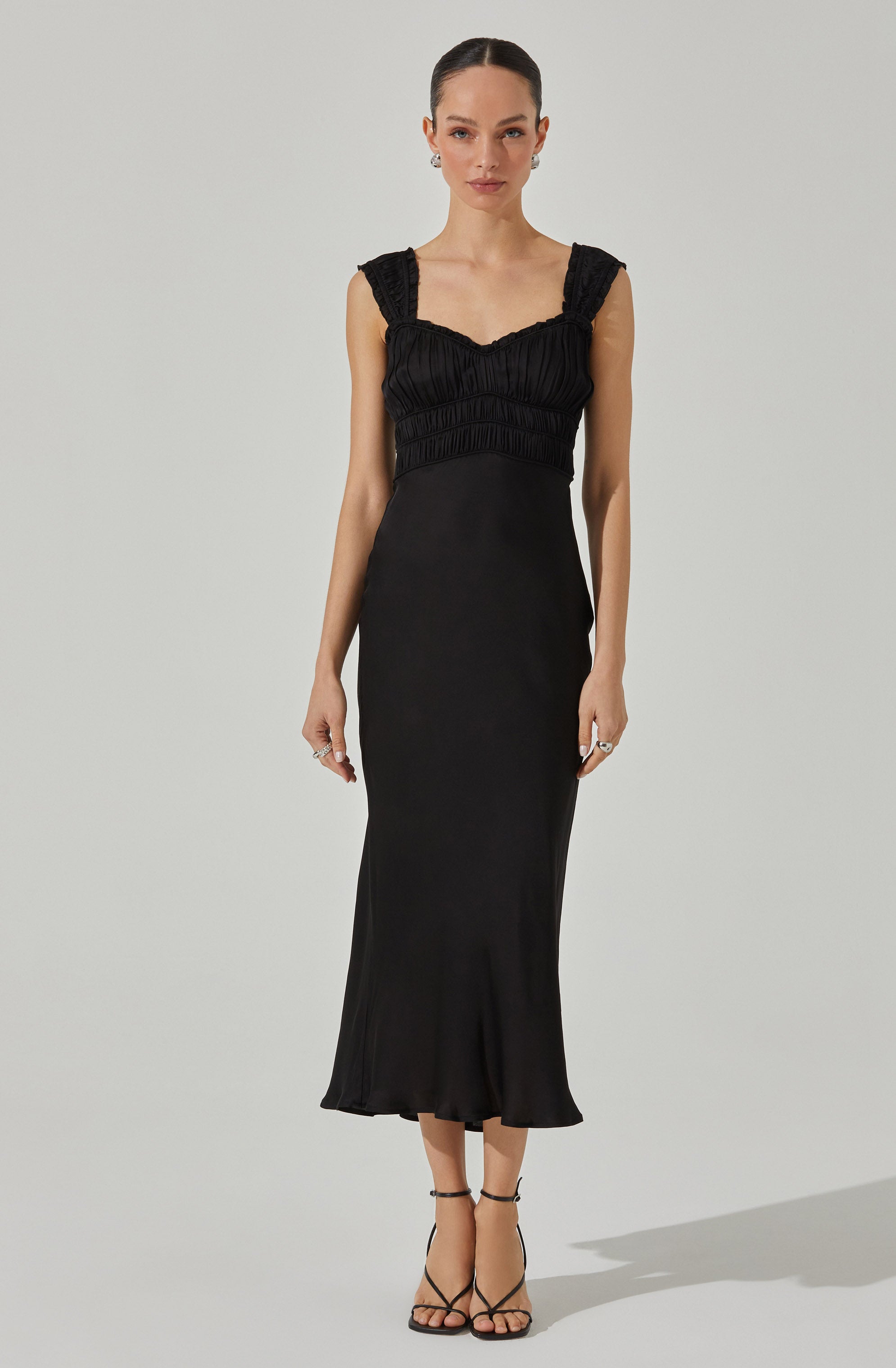 A woman is wearing a long, elegant black dress with a fitted bodice that features ruffled, textured shoulder straps and a ruched design across the chest. The dress has a smooth, flowing skirt that reaches mid-calf, with a subtle flare at the hem. She pairs the dress with black strappy high-heeled sandals.