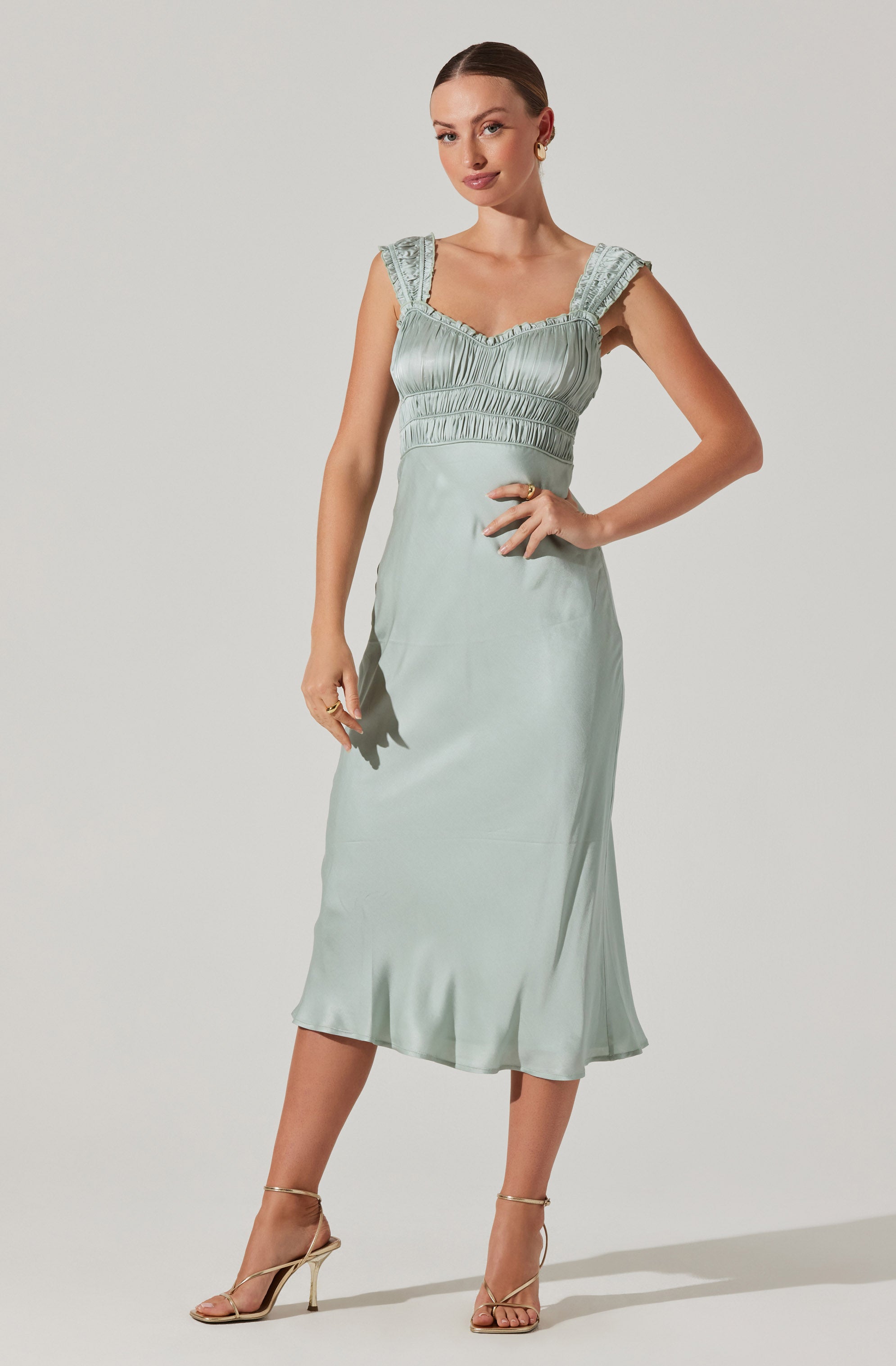 A woman is wearing a light mint green satin dress with a fitted bodice featuring gathered fabric detail and ruffled shoulder straps. The dress has a high waist and falls mid-calf length with a slight flare at the hem. She is also wearing strappy high-heeled sandals in a metallic gold color.