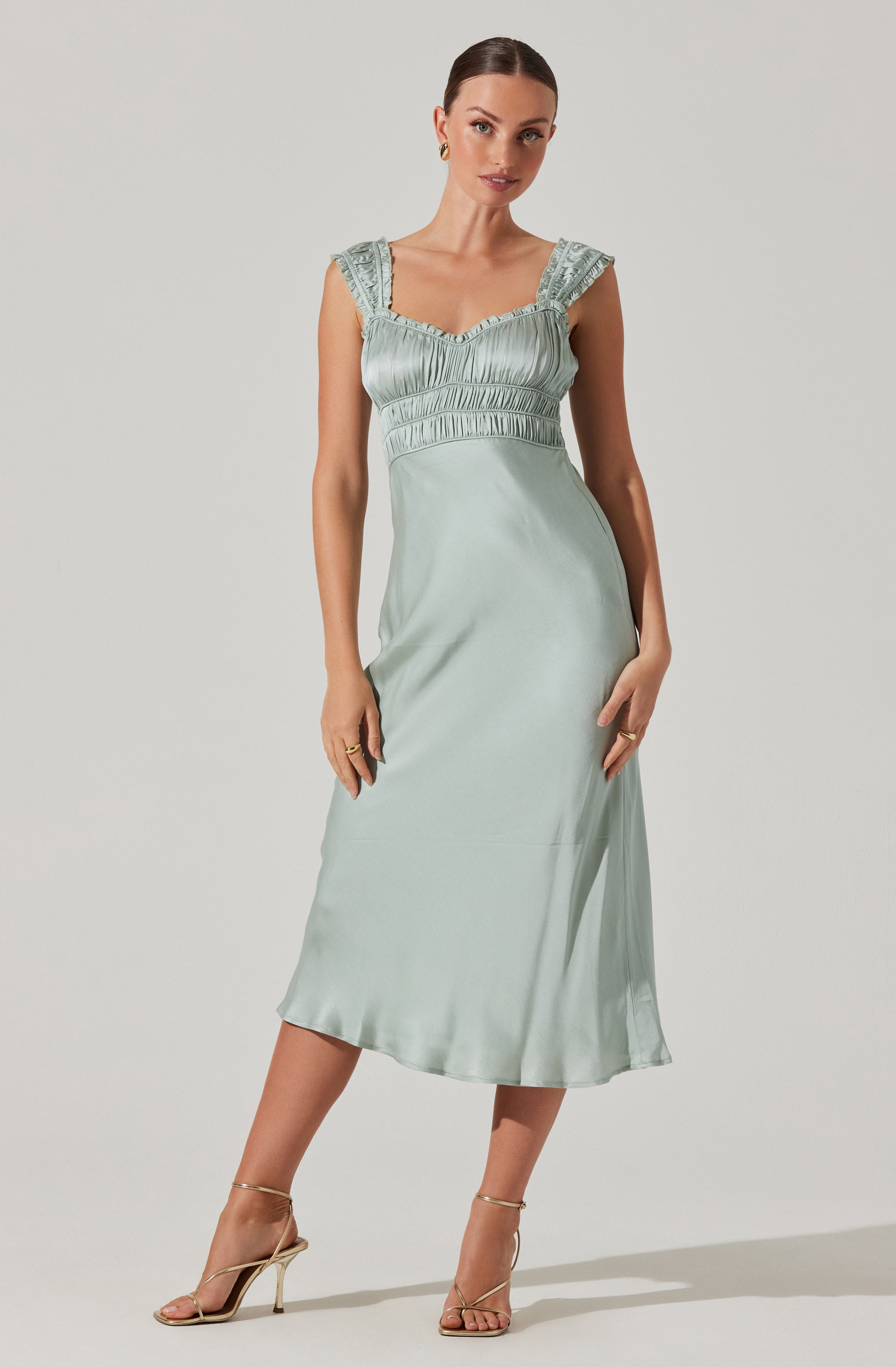 A woman is wearing a light green satin dress with ruffled straps and a smocked bodice. The dress has a fitted waist and flows down to a midi length. She pairs the dress with gold high-heeled sandals.