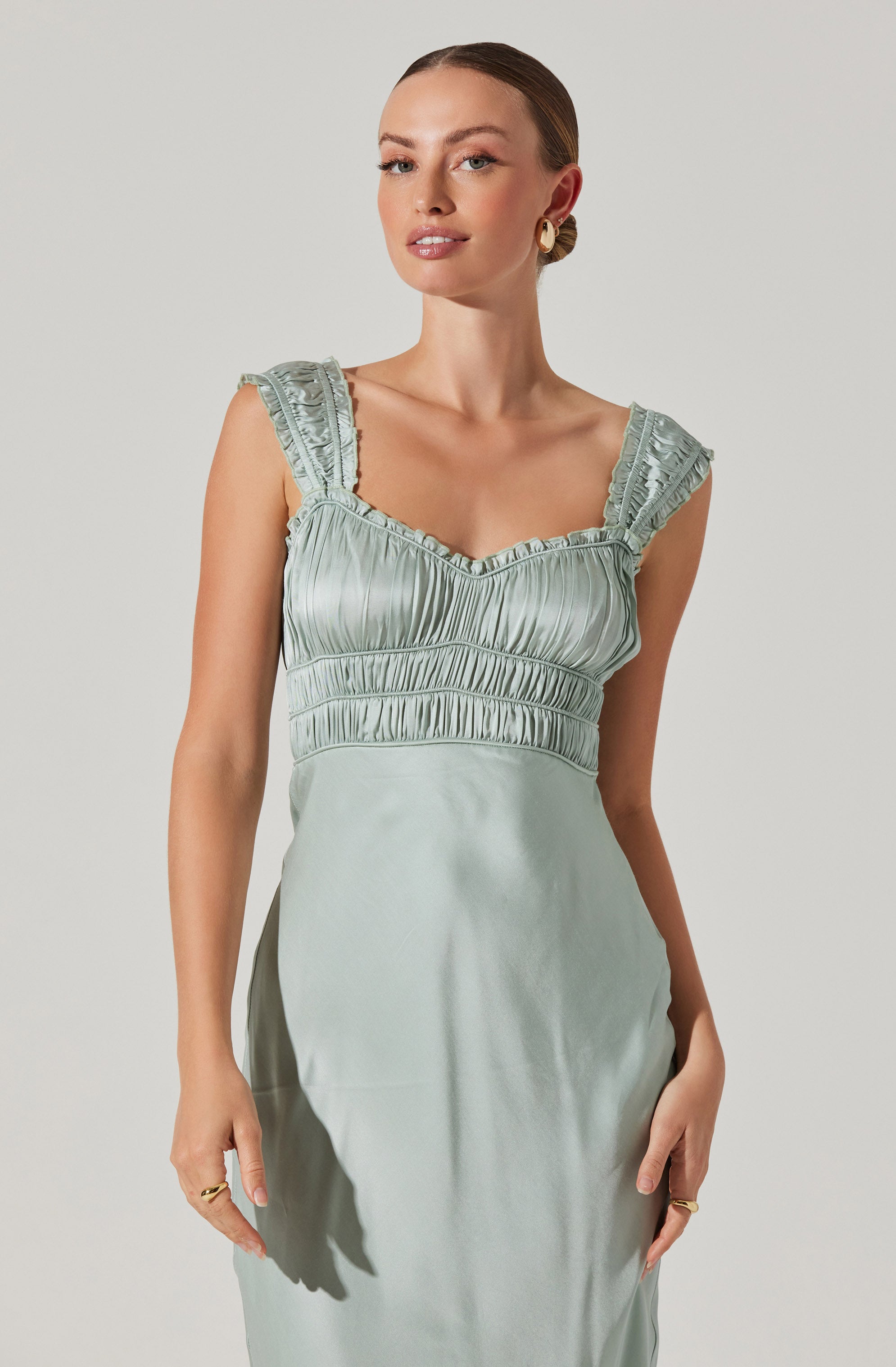 A woman is wearing a sleeveless, light green satin dress with ruffled shoulder straps, a ruched bodice with vertical stripes, and a smooth skirt.